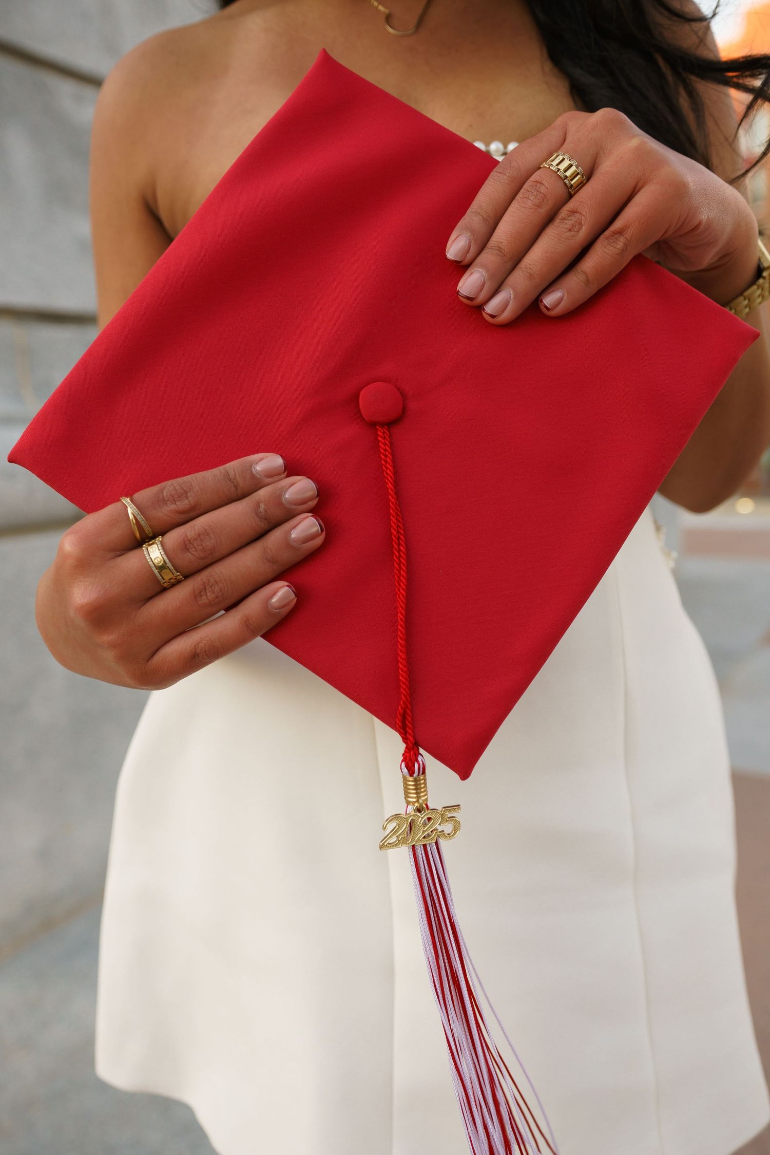 A woman in a white dress holds a red graduation cap with a gold 2025 tassel charm, wearing gold rings.