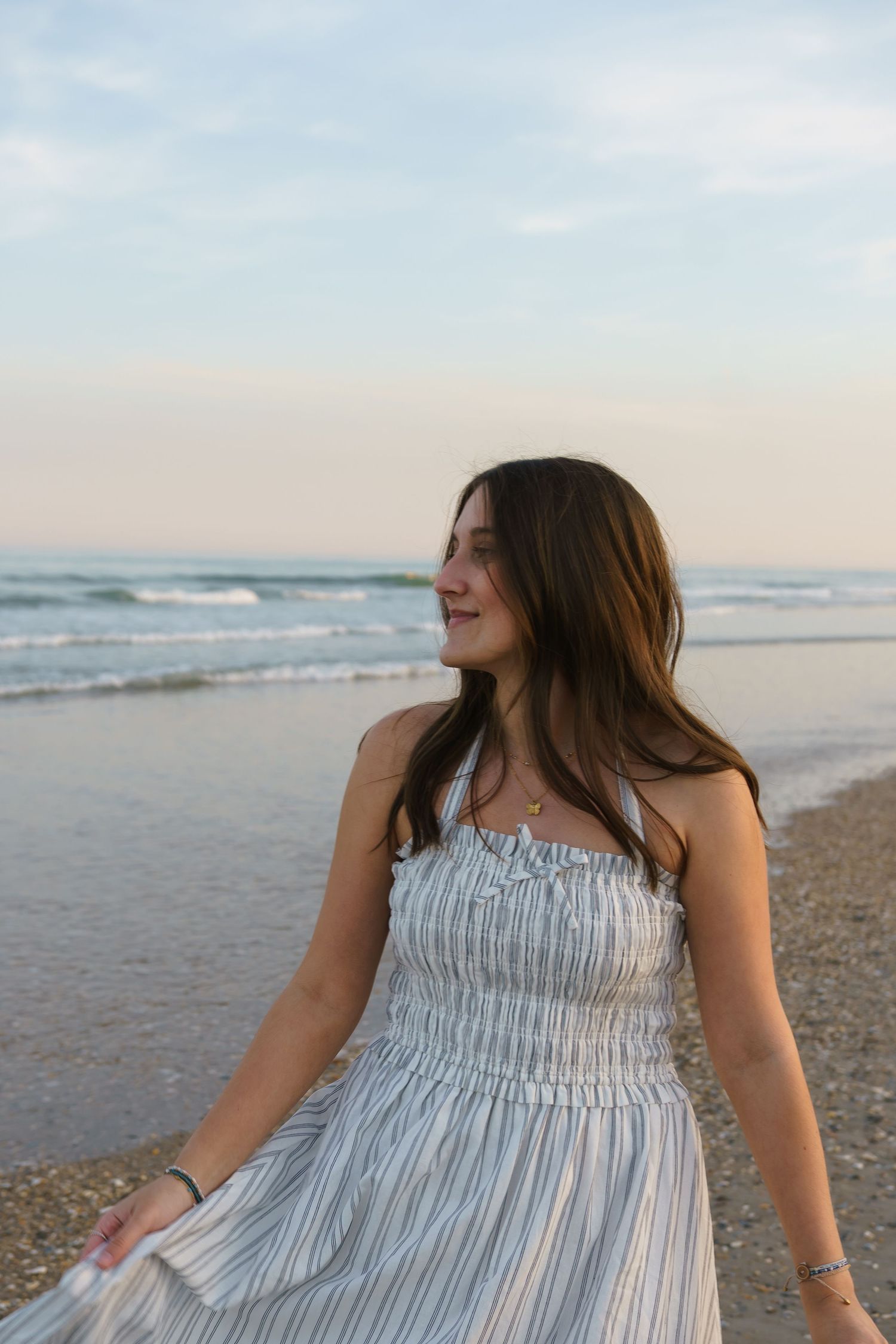 Young brunette woman in striped strapless maxi dress smiling on ocean beach at dusk with waves behind her.