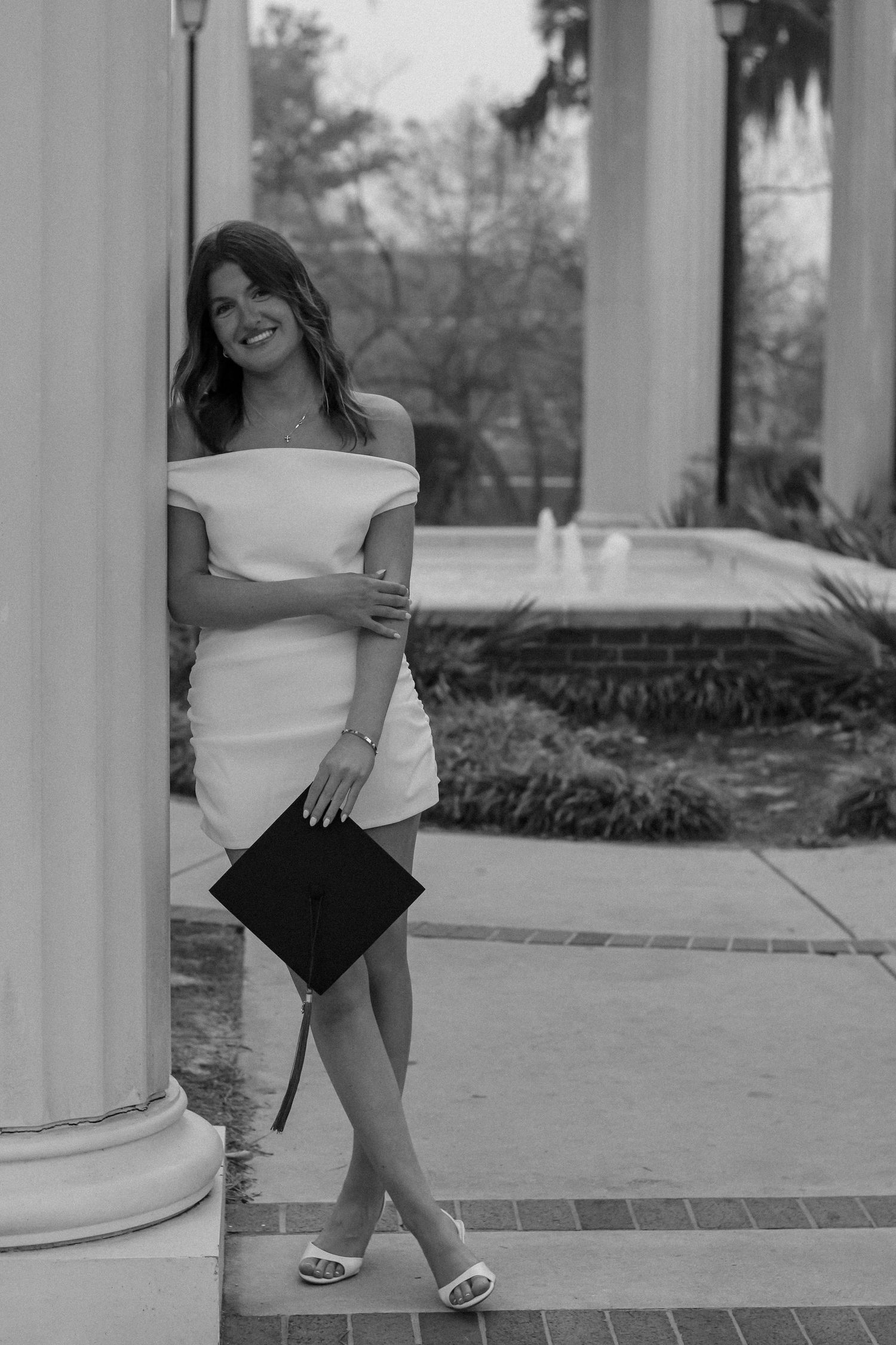 Black and white photo of graduate in white dress leaning against column with graduation cap.