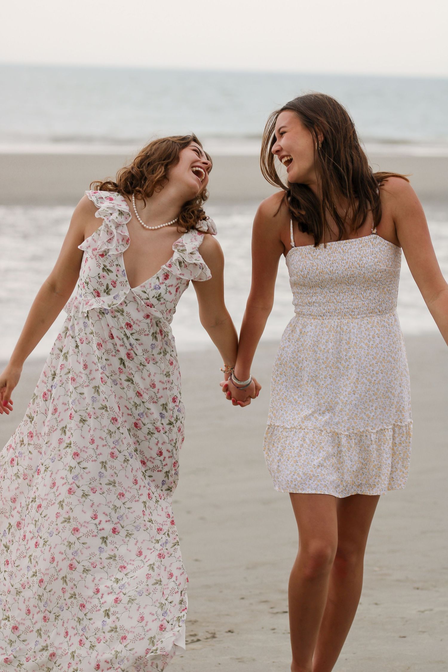 Two friends in white summer dresses share joyful moment while walking on beach.