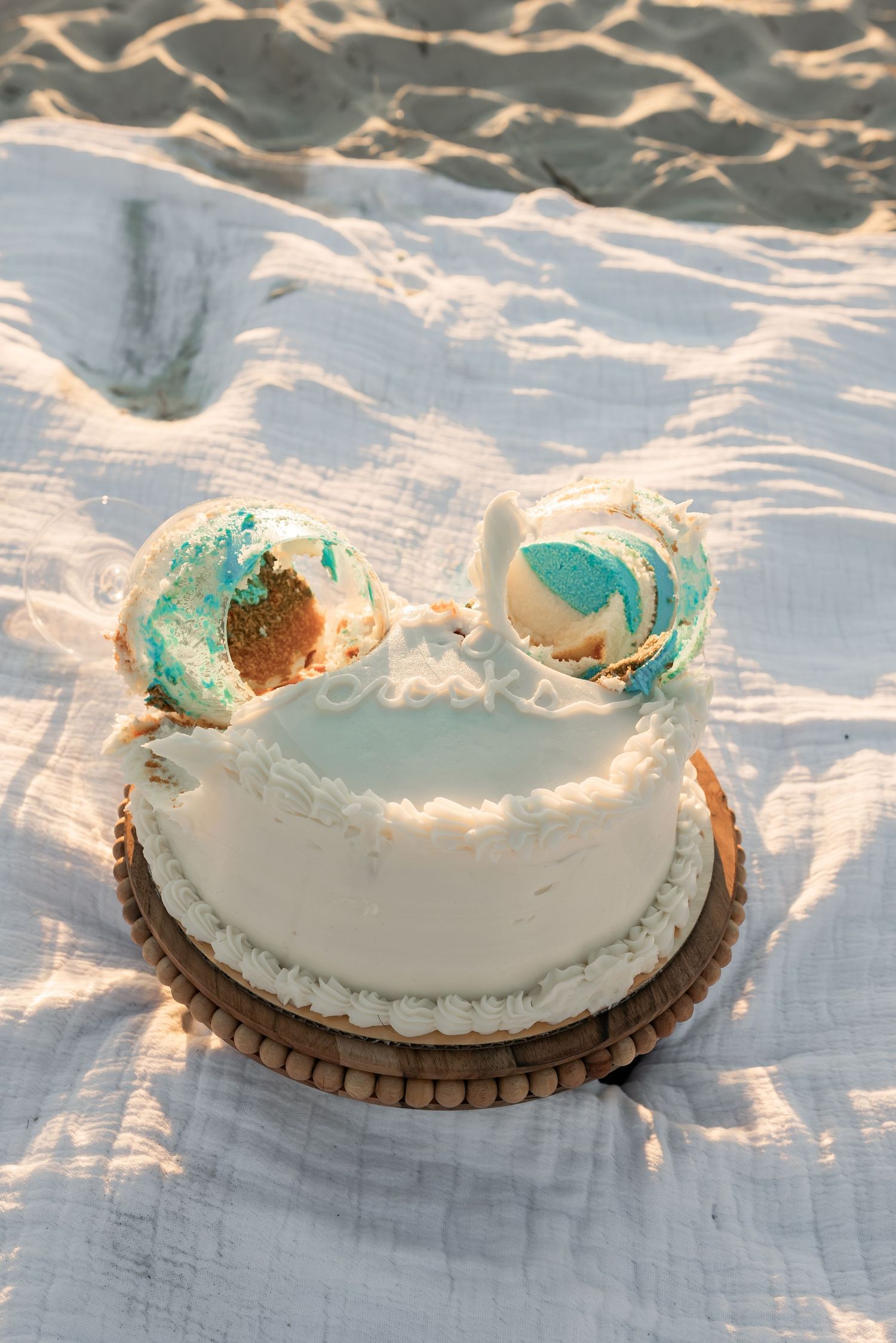 White cake with turquoise details on a wooden platter against a beach backdrop.