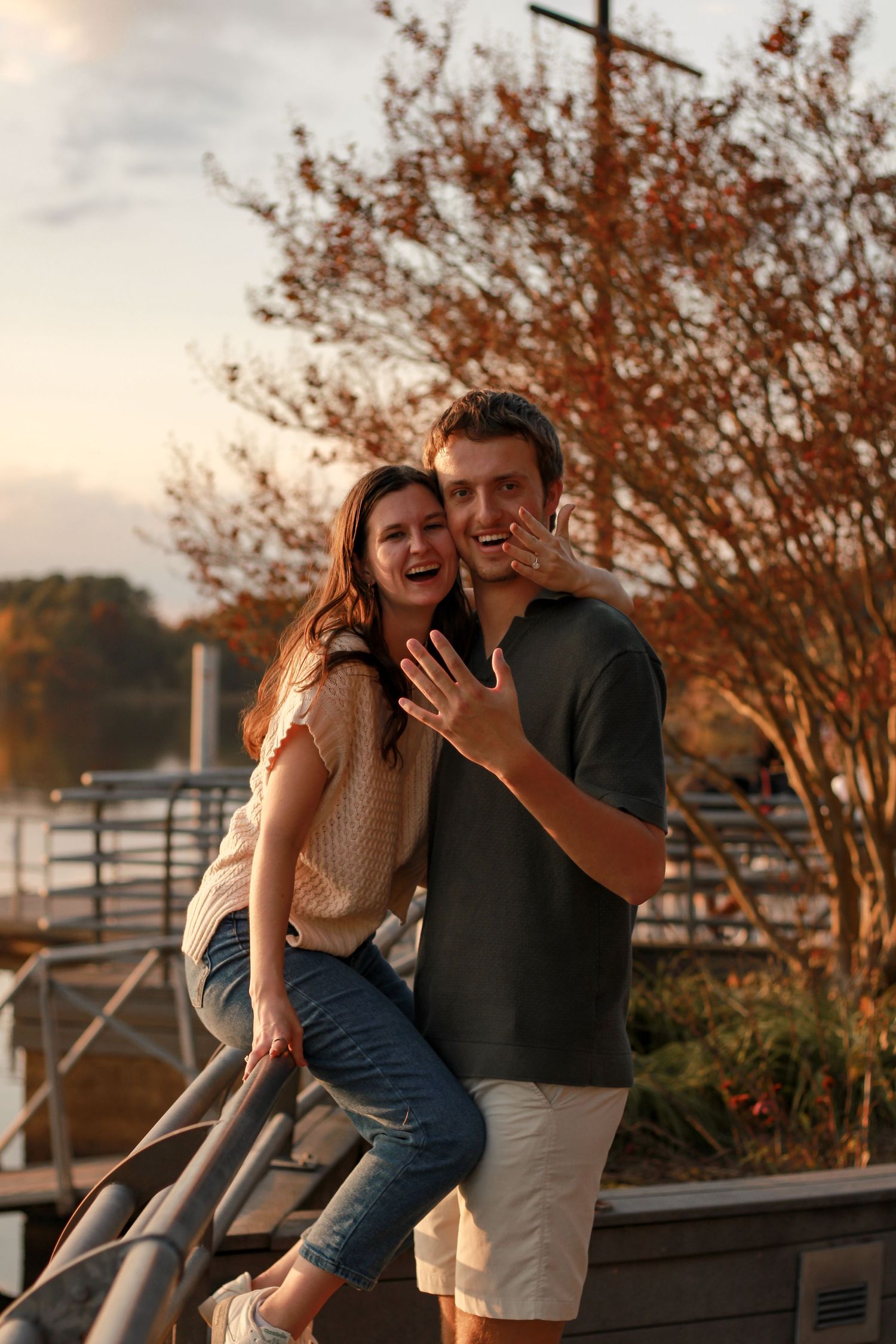 A couple shares an intimate moment at sunset on a waterfront deck with fall foliage in the background.