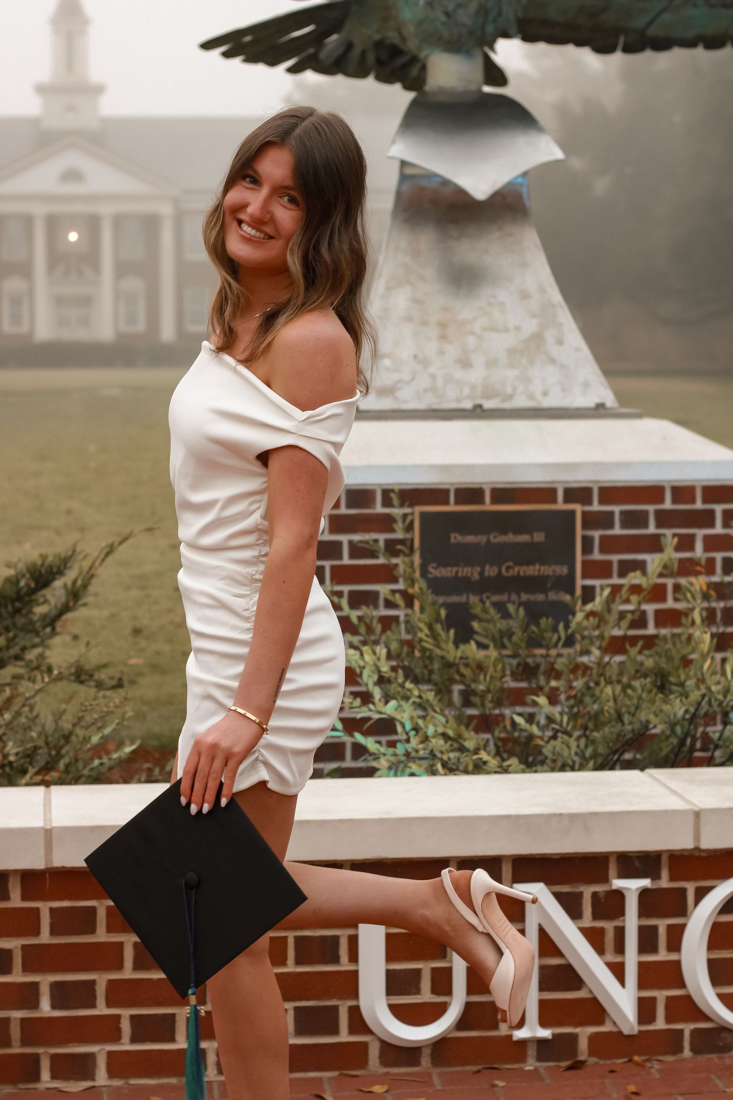 Graduate in white dress holding cap by UNCW campus statue.