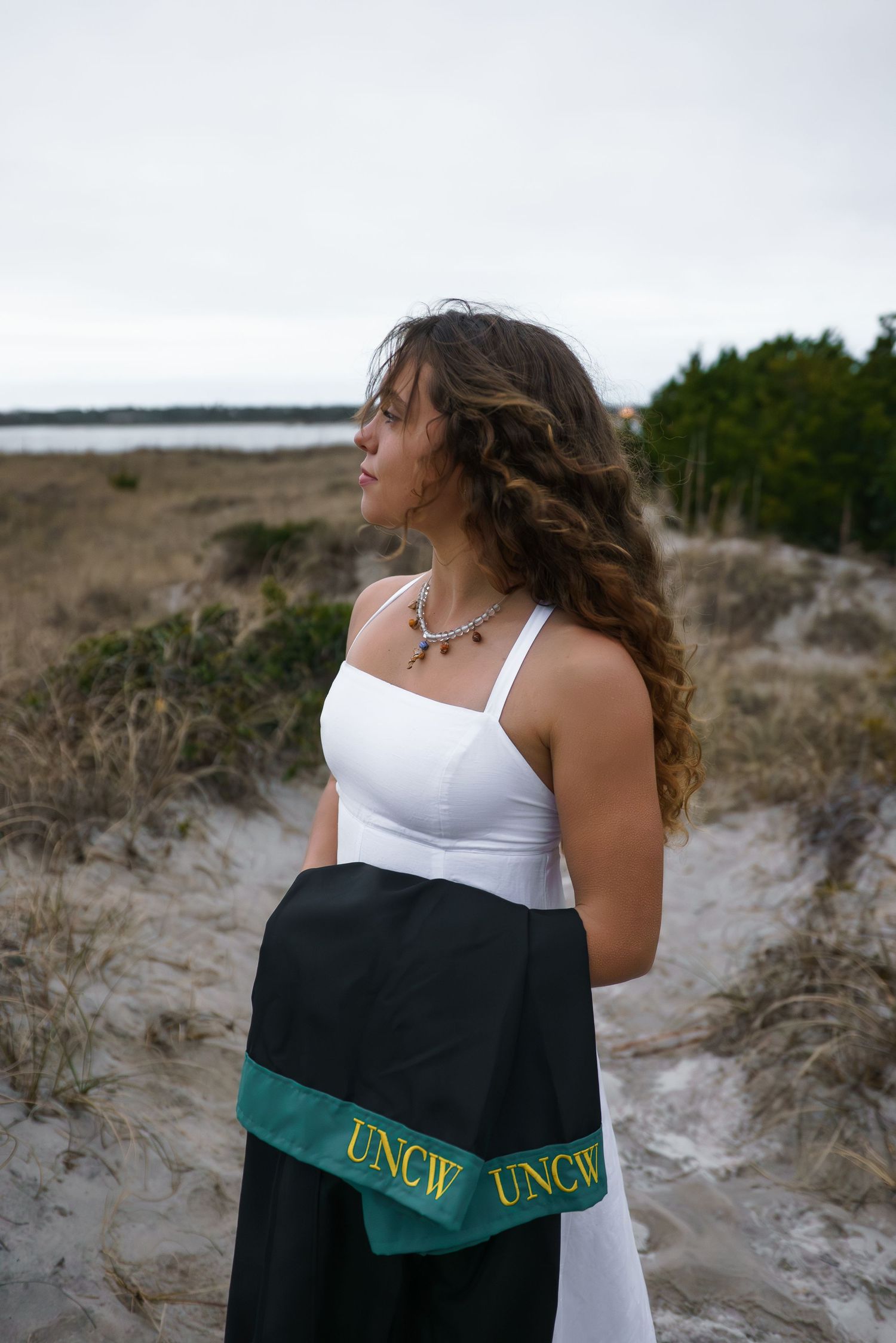 Graduate wearing UNCW gown poses against sandy dunes during a photo session.