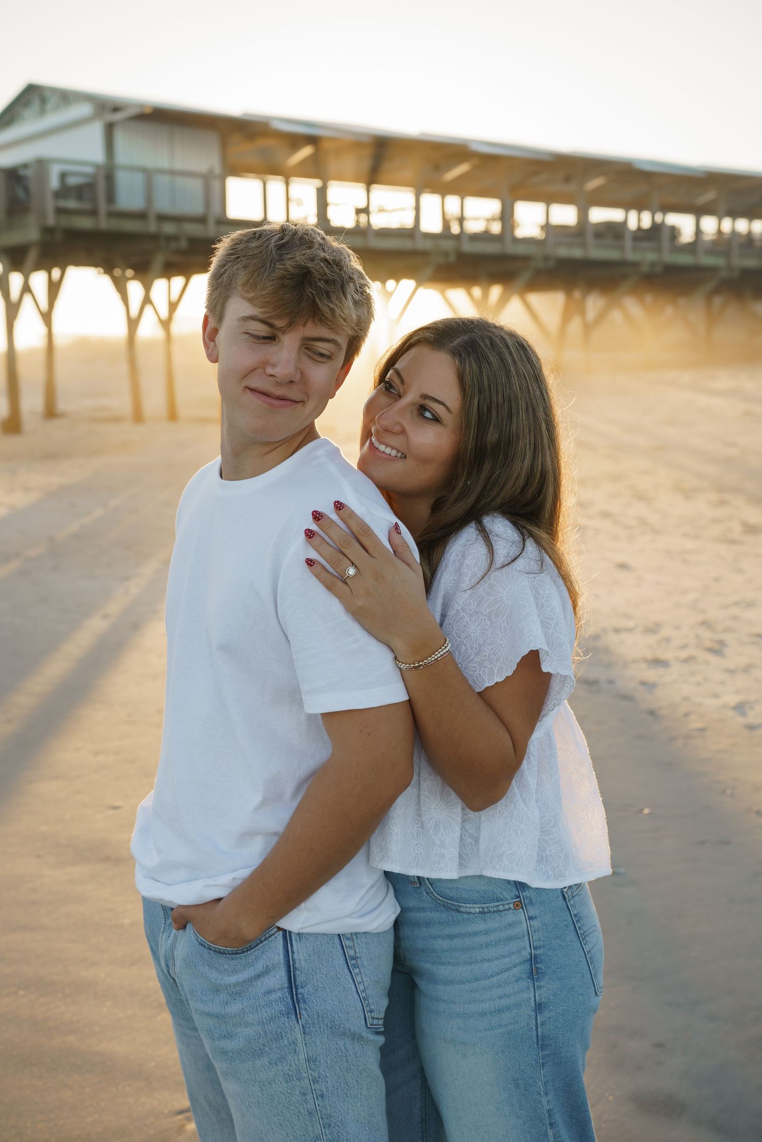 A couple posing together on the beach with a pier structure in the background at sunset.