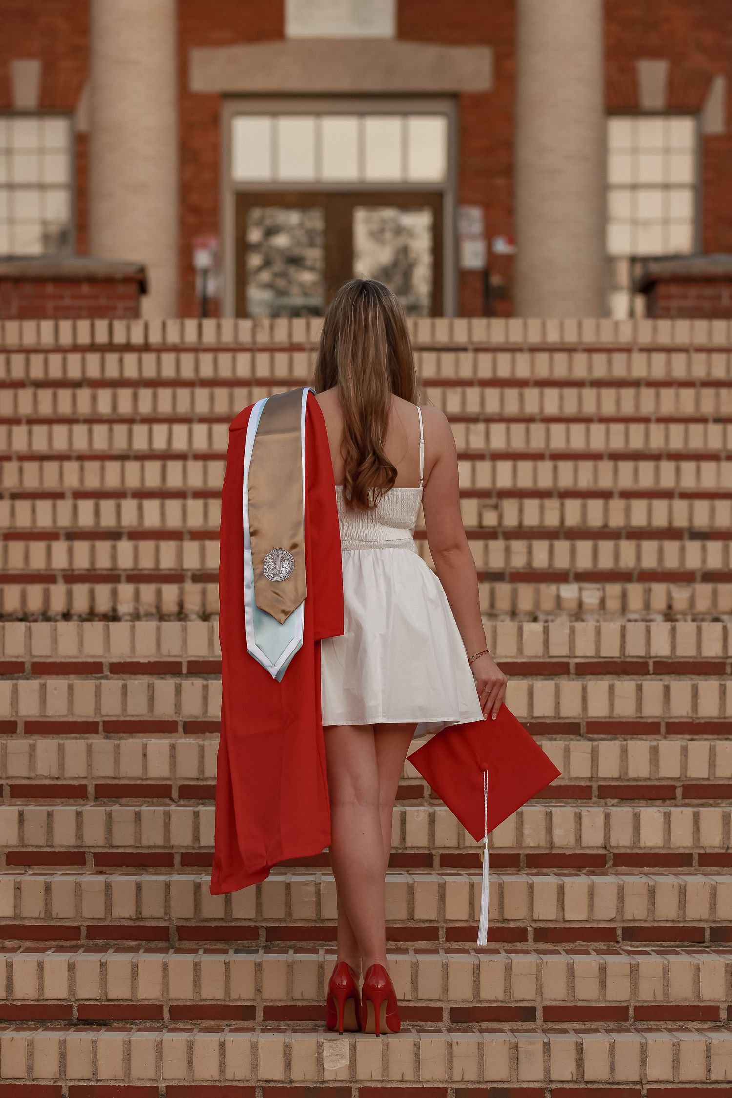 Red graduation cap and gown displayed on brick steps of academic building.