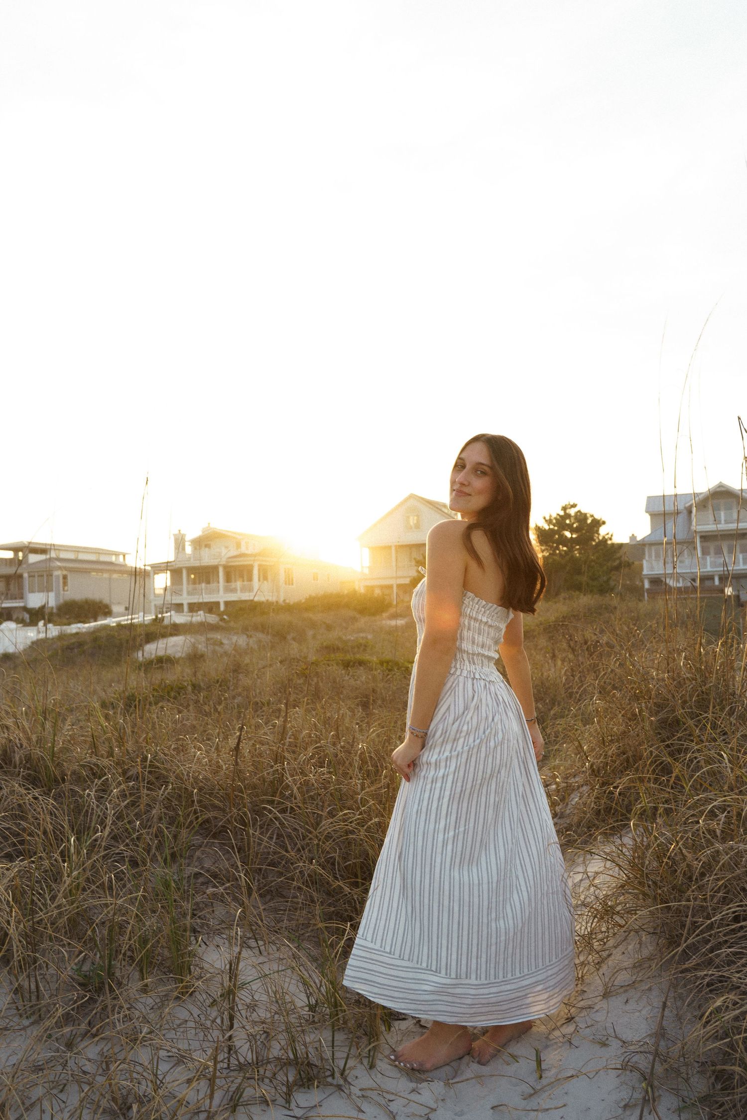 Young woman in striped sundress standing on beach sand dunes at golden sunset with coastal houses in background.