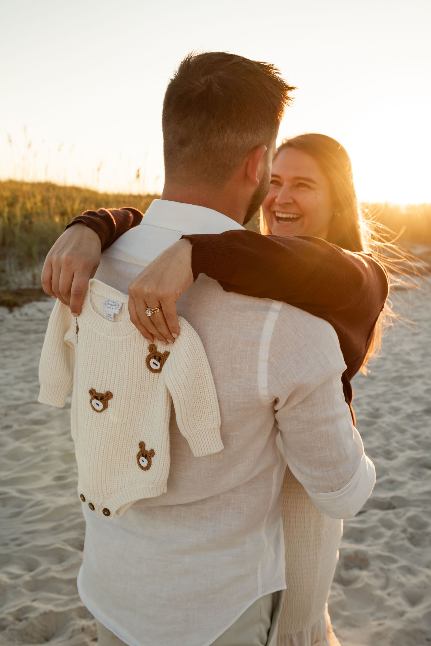 Couple embracing on beach at sunset while holding a small white onesie.