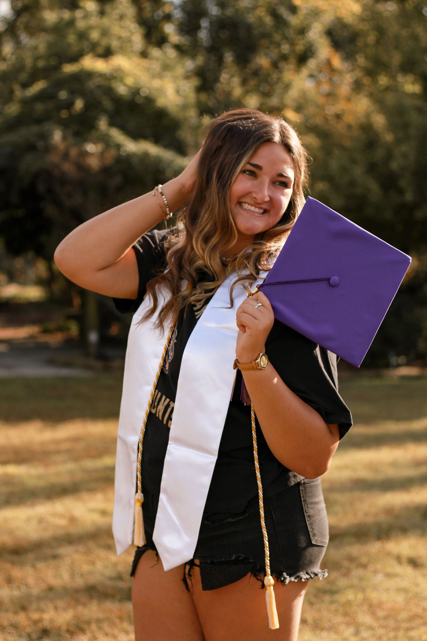 Graduate poses outdoors holding purple graduation cap while wearing graduation stole.