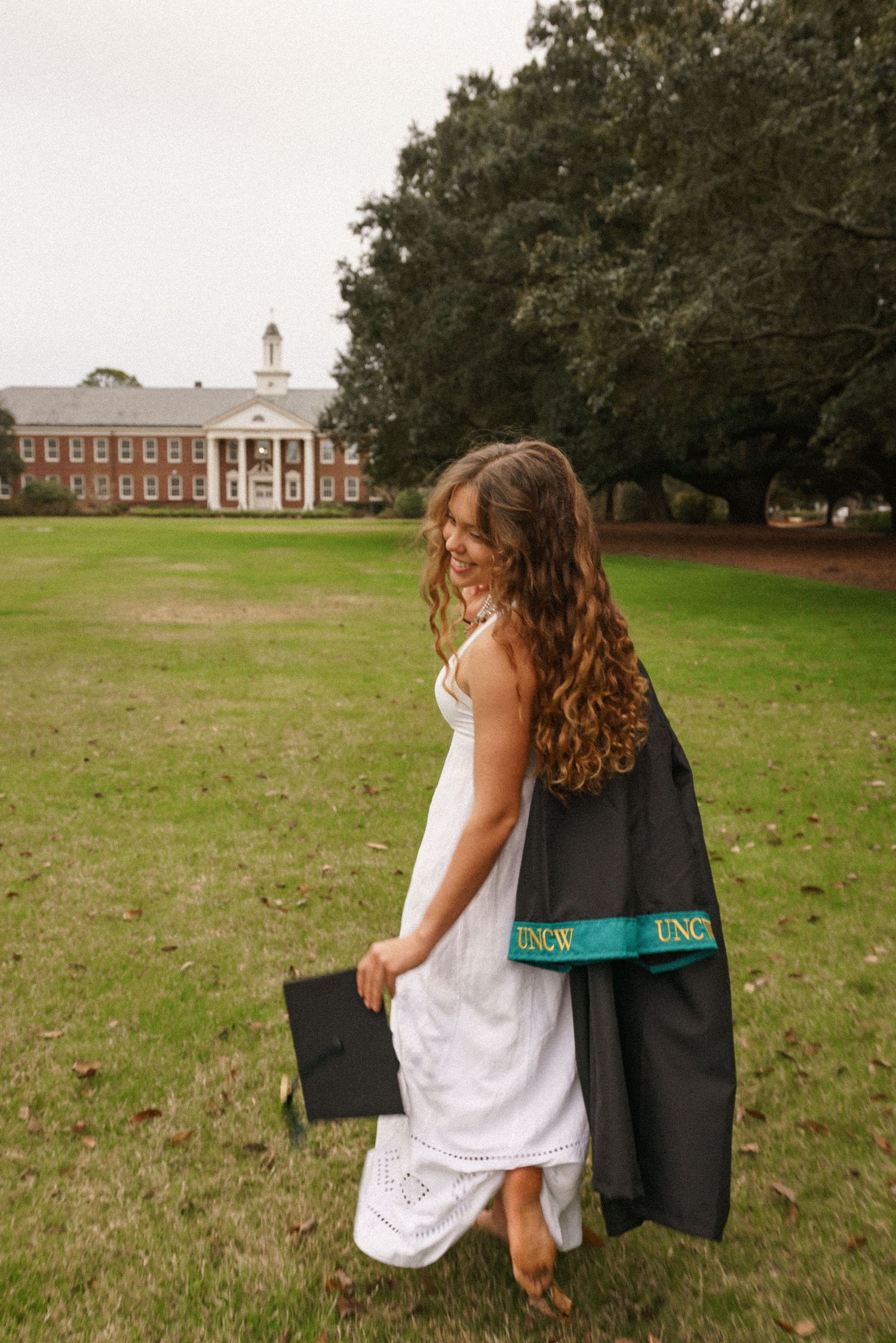 Graduate in white dress runs across UNCW lawn with academic building in background.