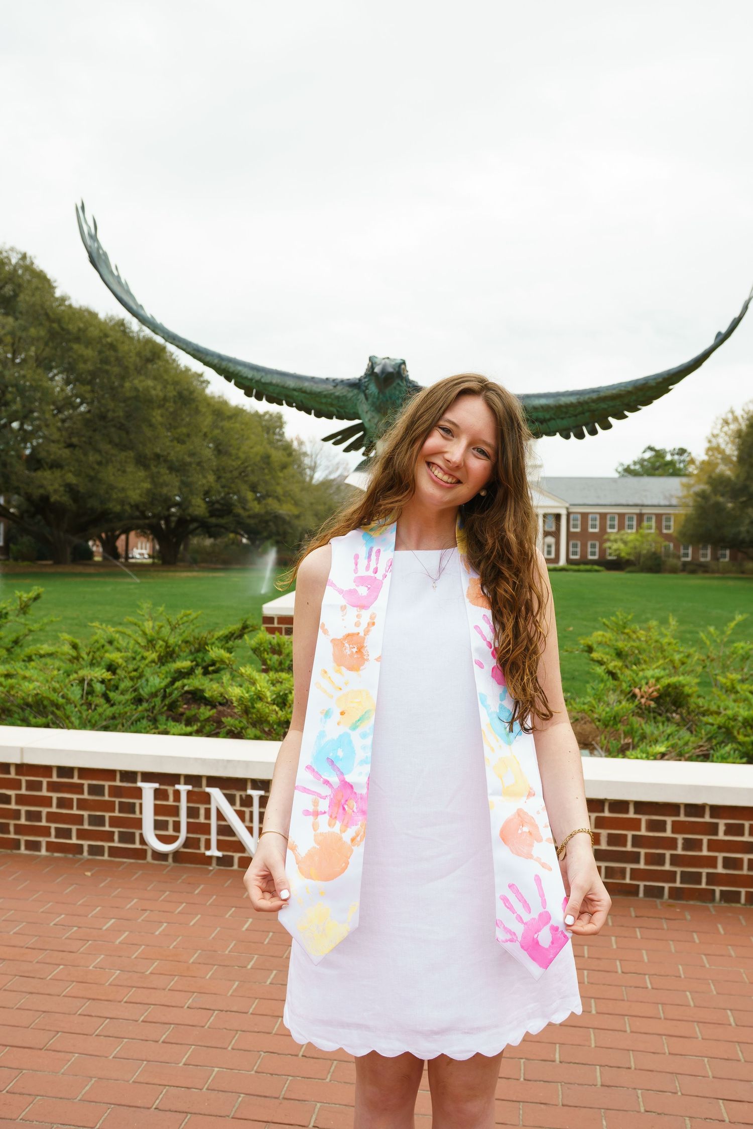 Smiling graduate in white dress with colorful handprint sash poses before bronze eagle statue on brick campus plaza.