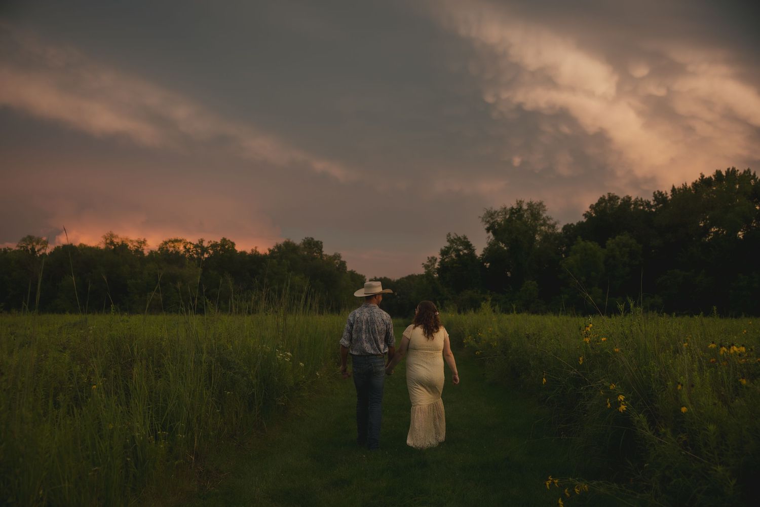 A couple walks together through a serene meadow at sunset with tall grass and trees silhouetted against the dimming sky.