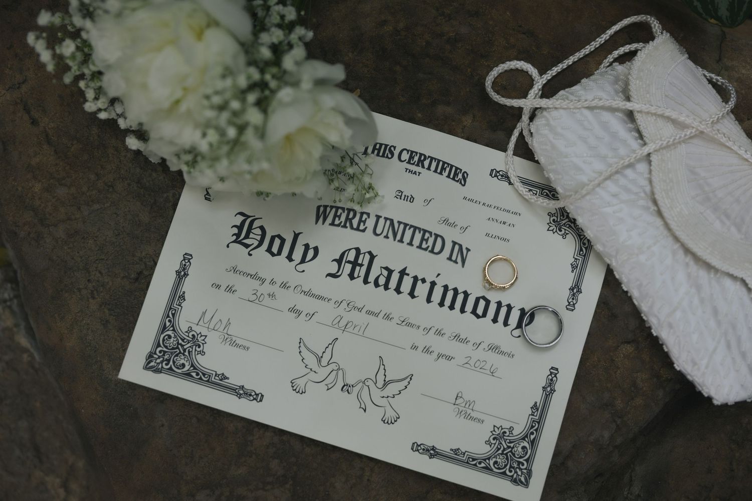 Holy Matrimony marriage certificate with wedding rings, white flowers, and rope on stone background.
