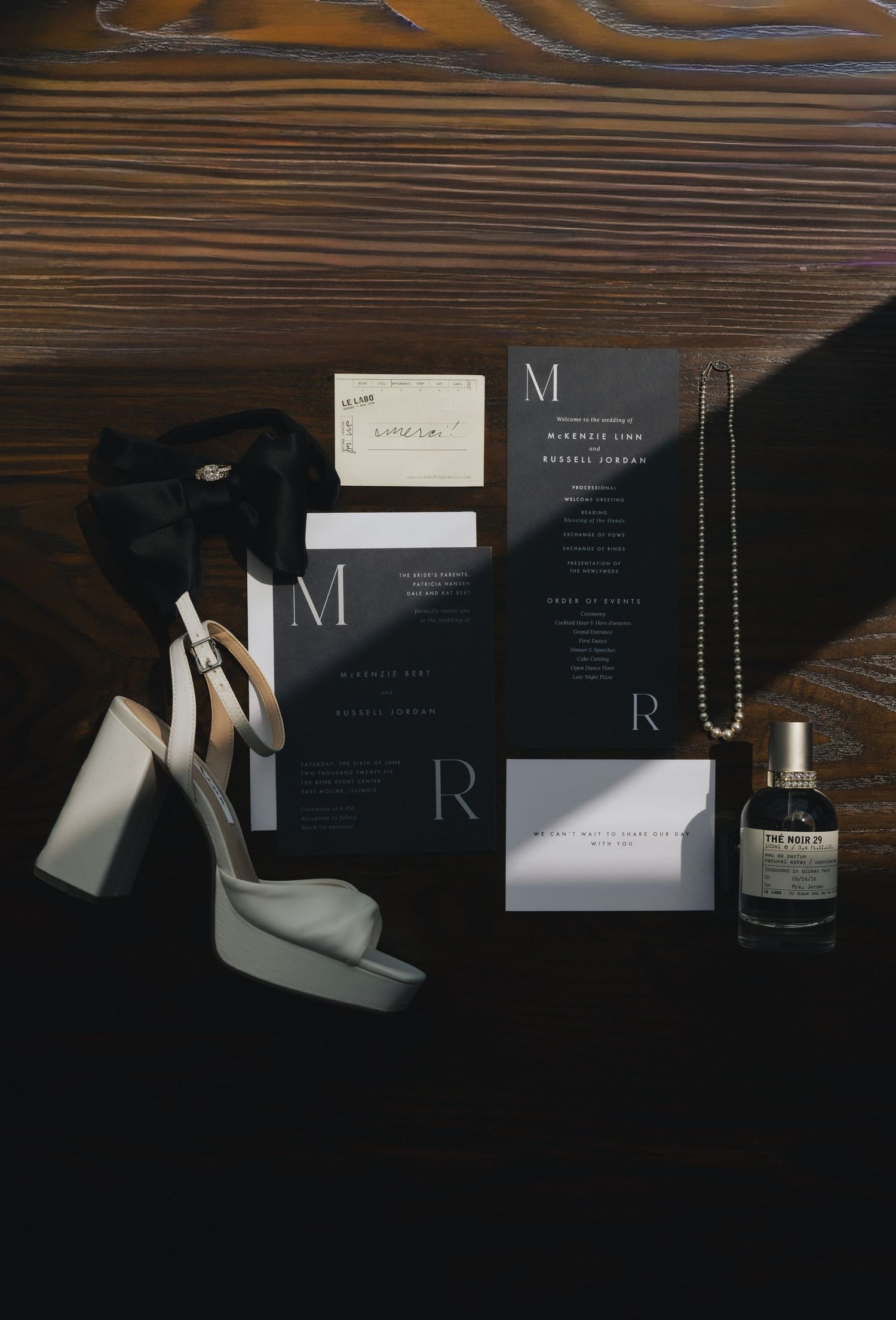 Elegant dark wedding invitation suite with monogram letters M and R, white heels, pearl necklace, and perfume bottle on wood.