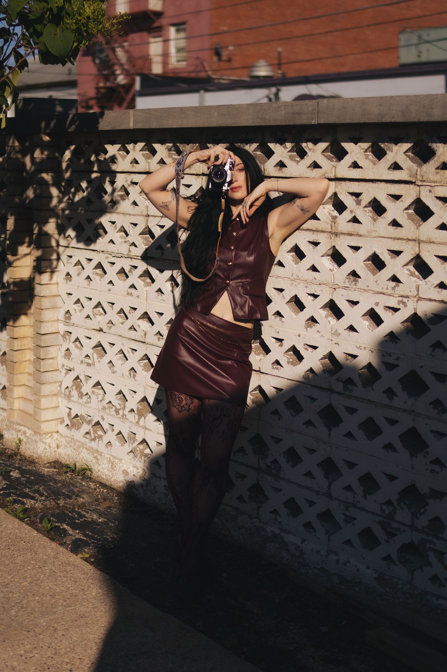Woman in burgundy leather co-ord holds vintage camera outdoors against a white lattice fence in sunlight.