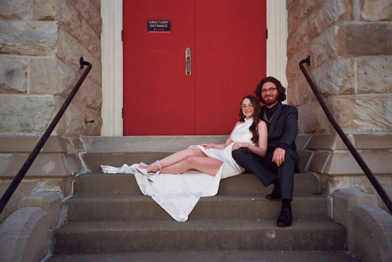 Couple in wedding attire sitting on stone steps before a red door, bride in white dress, groom in dark suit.