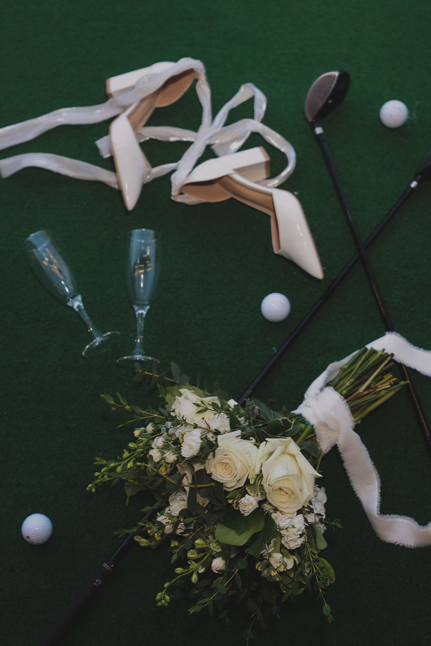 Wedding details with ballet shoes, floral bouquet, champagne glasses and golf clubs arranged on dark green background.