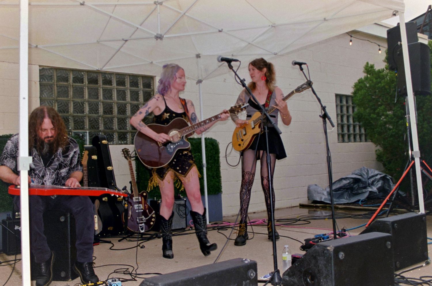 Three musicians performing on stage under a tent, two women playing guitars and a man at a keyboard.