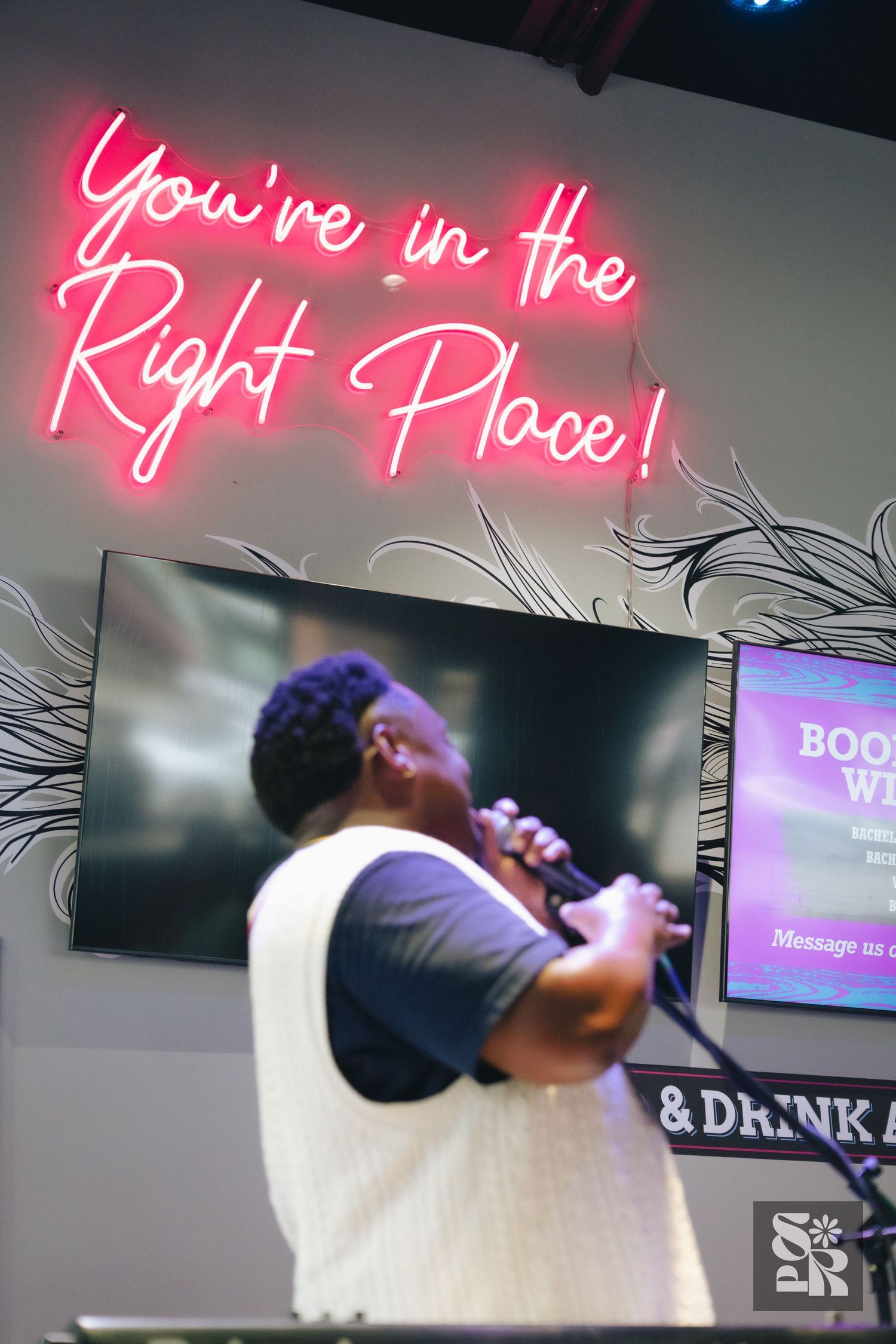 Person singing into microphone under pink neon sign reading You're in the Right Place at a lively venue event.