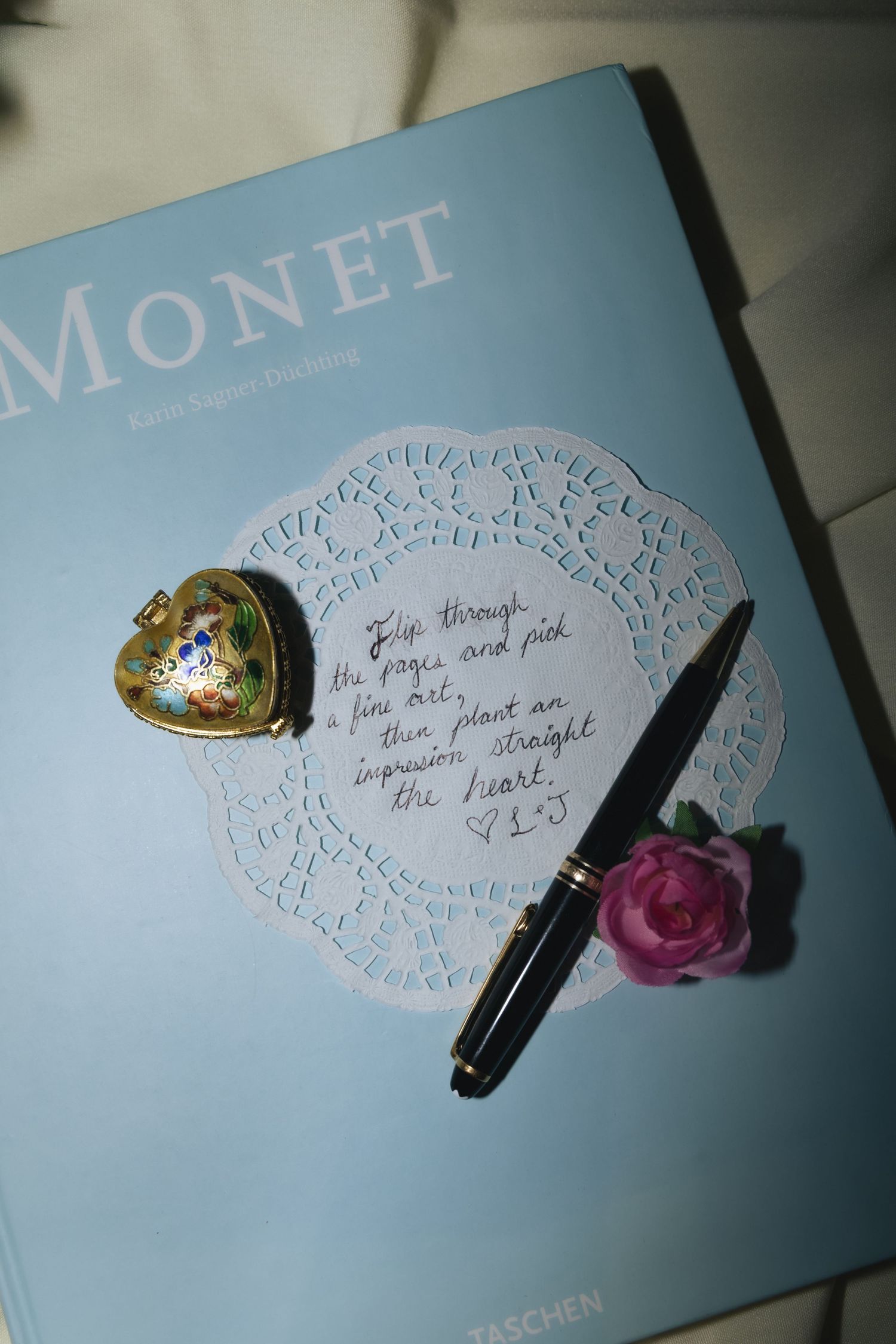 Monet Taschen book with a heart locket, pink rose, pen, and handwritten note on a lace doily.