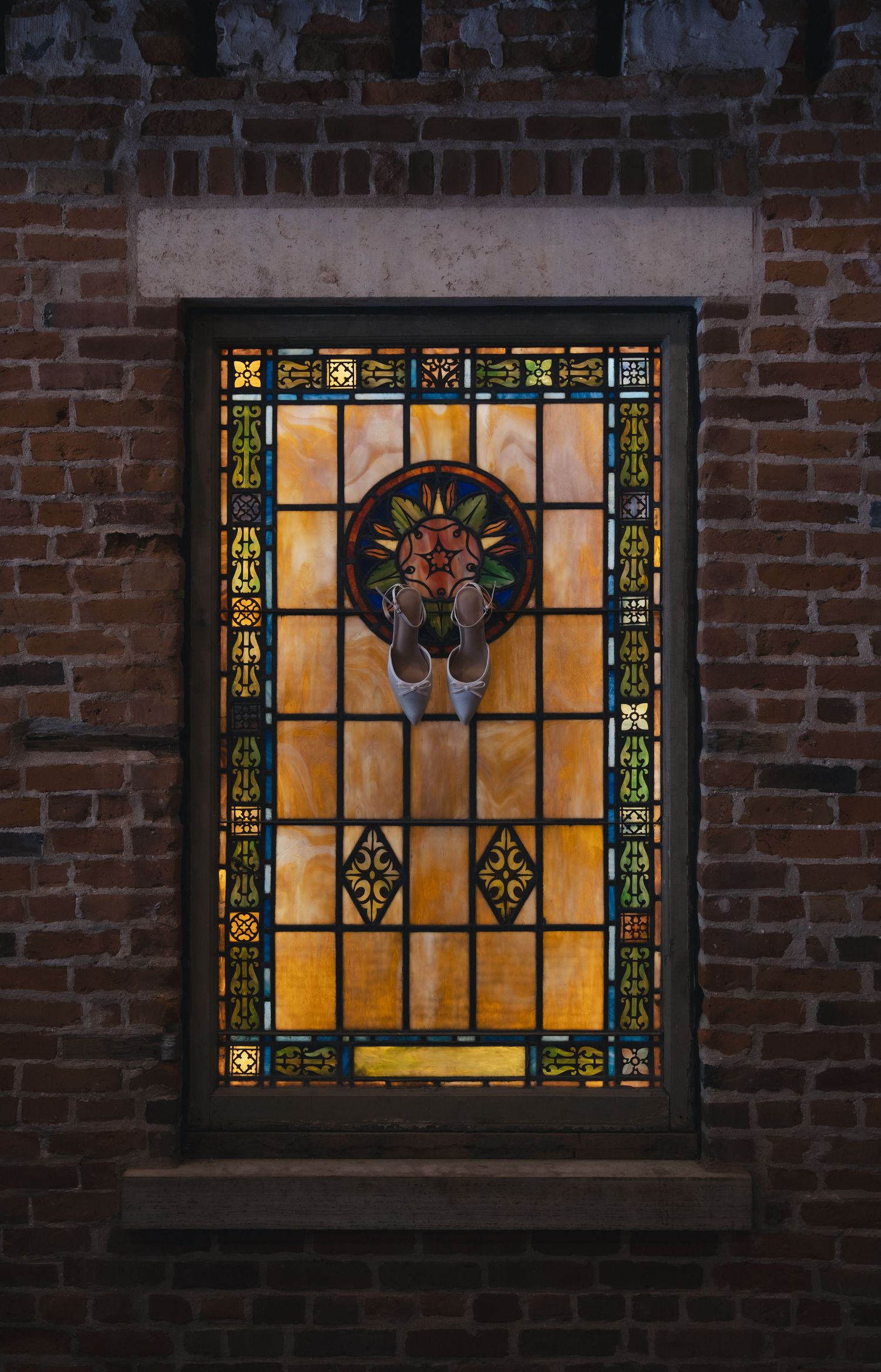 Ornate stained glass window with amber tones and floral medallion set in a rustic red brick wall.