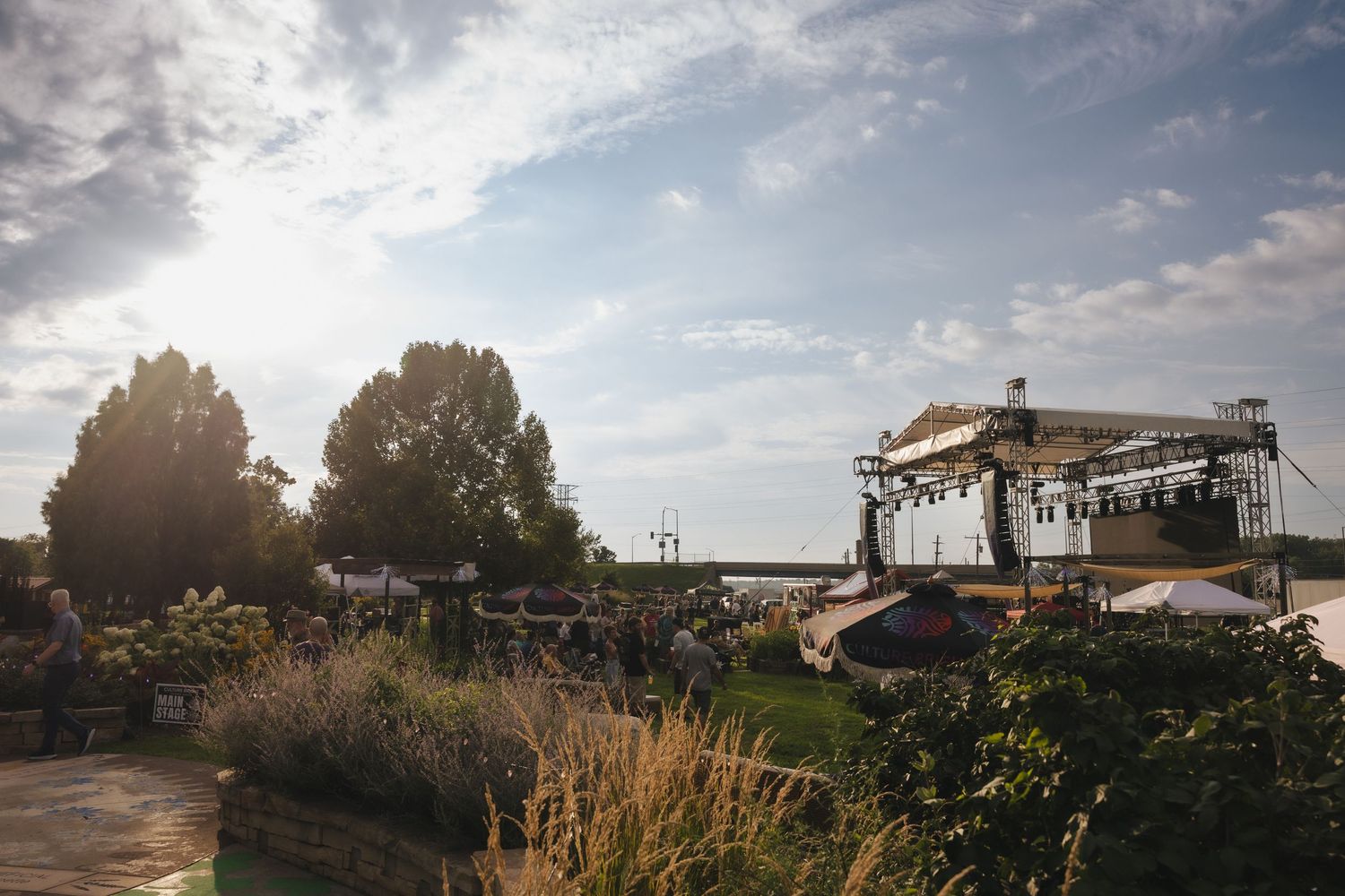 Outdoor concert stage with scaffolding lighting rig set against a scenic waterfront park with lush greenery and dramatic sky.