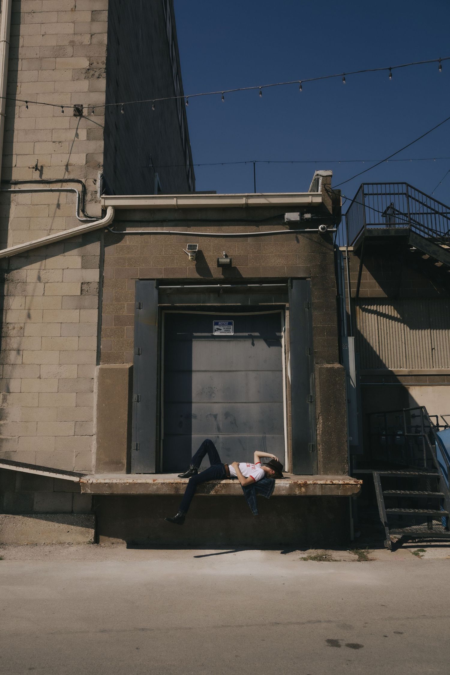 Person lying on a concrete ledge outside an urban industrial building with metal stairs and string lights under a clear blue sky.