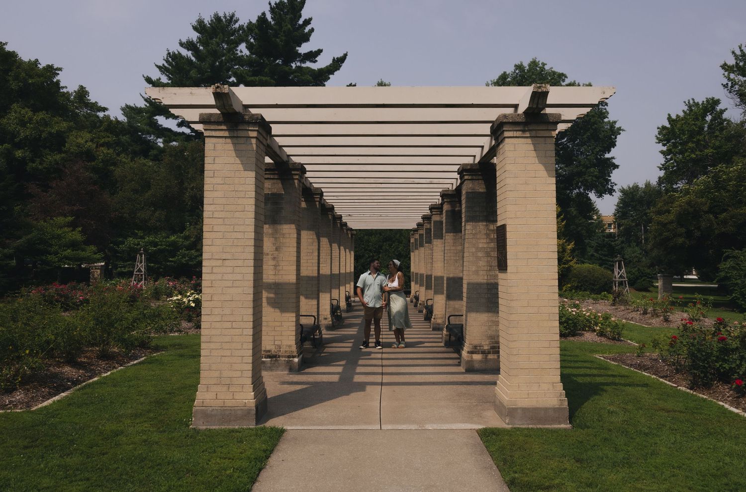 Couple walking through a pergola garden pathway with stone columns and white lattice roof surrounded by lush greenery.
