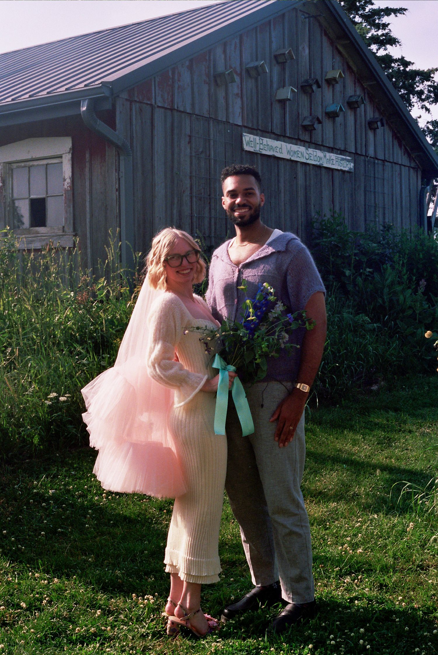 A couple stands in front of a rustic barn, wearing formal attire with the bride in a short pink tulle dress.