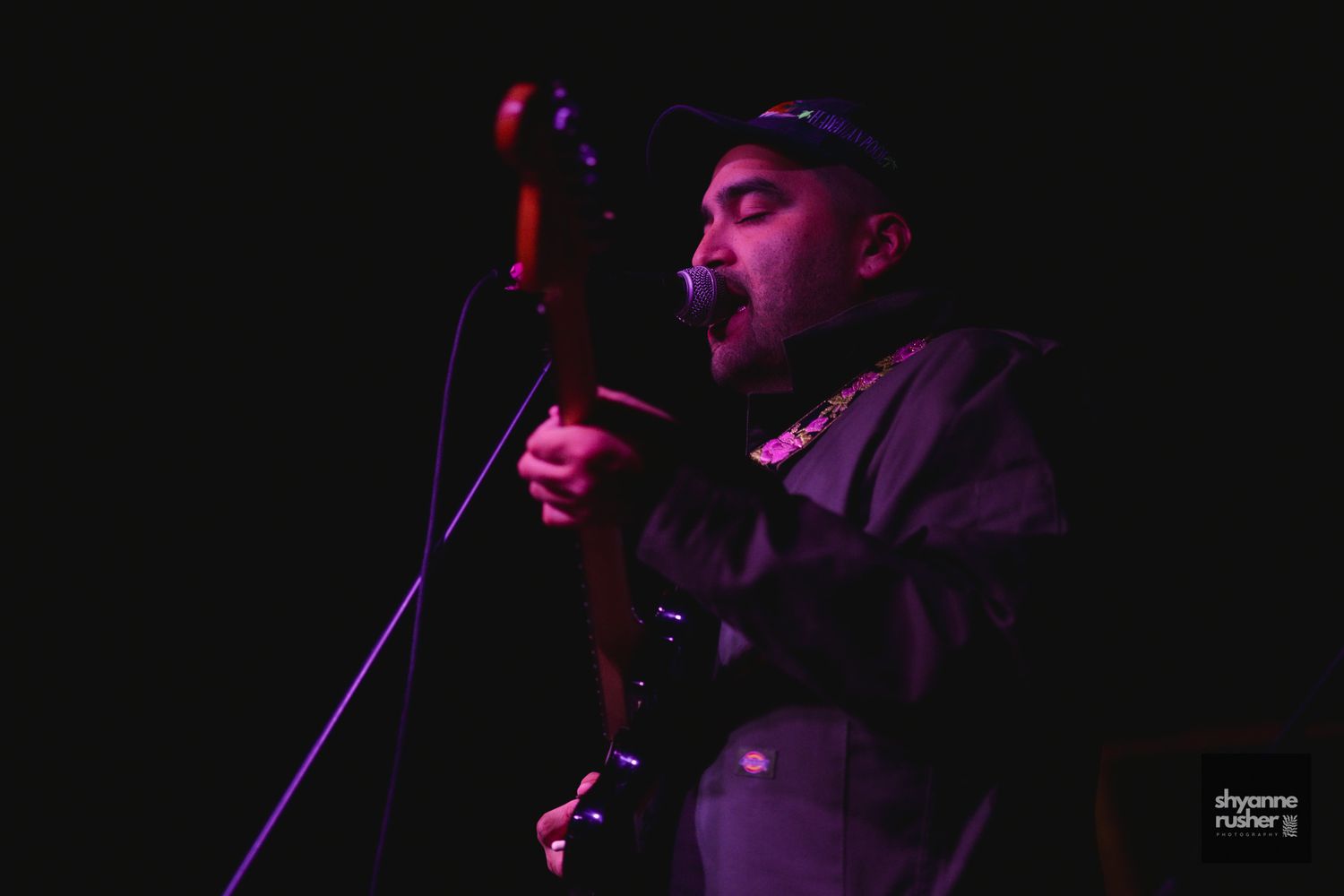 A musician performs on stage in low purple lighting while singing into a microphone and playing guitar.