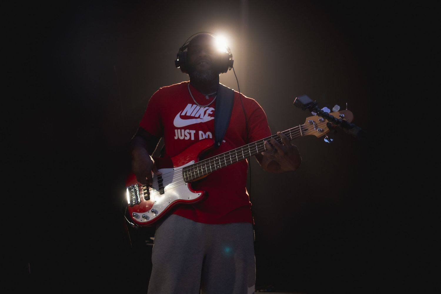 Bass guitarist in a red Nike shirt plays under dramatic spotlight on dark stage.
