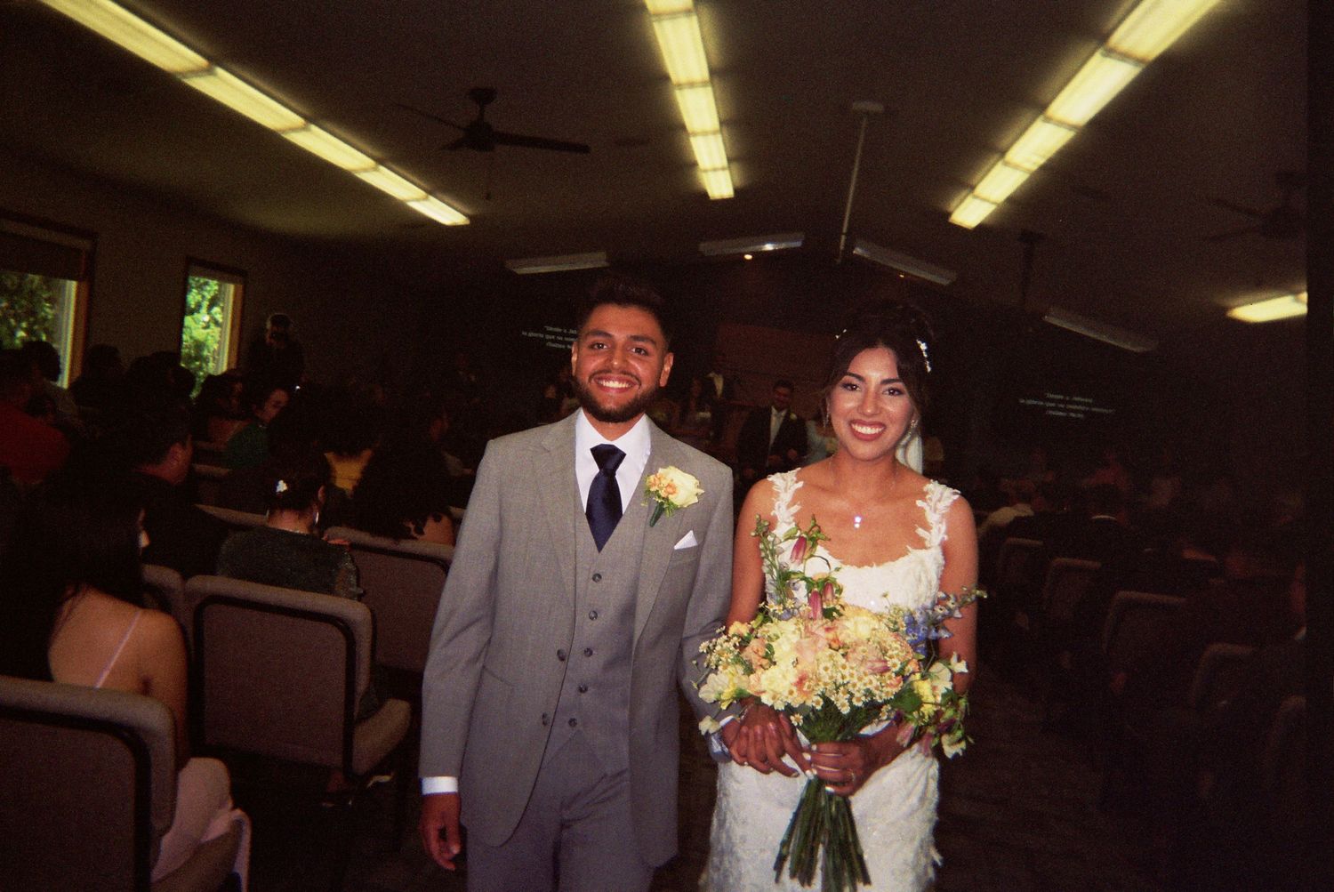 A wedding ceremony takes place in a simple indoor venue with fluorescent lighting overhead.
