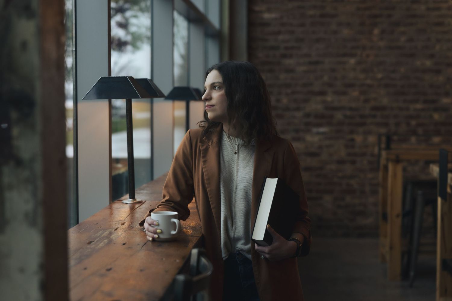 Woman with long hair holding book and coffee cup gazing out window in cafe.