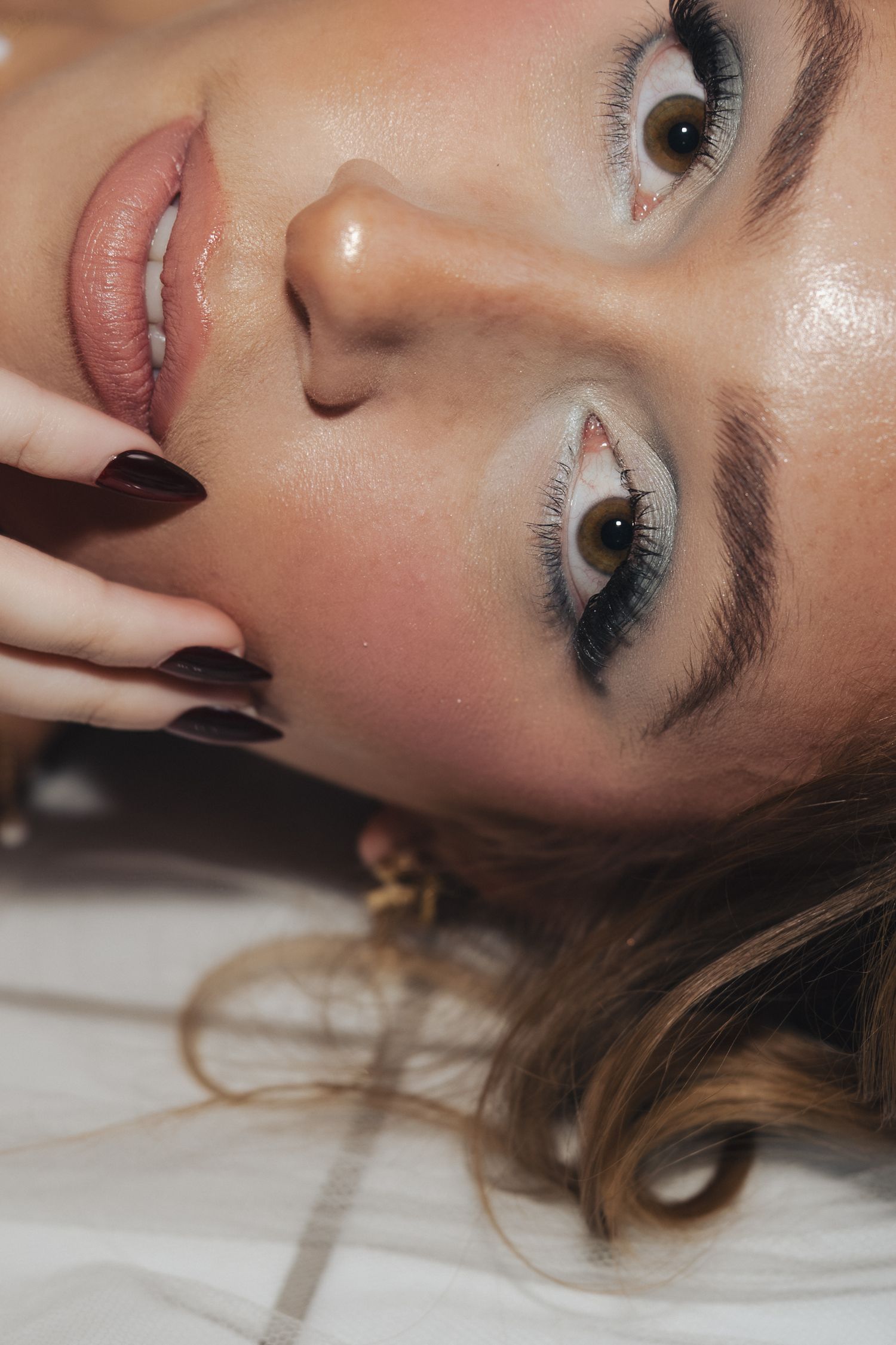 Close-up beauty portrait of person with smokey eye makeup and glossy lips laying down against white background.
