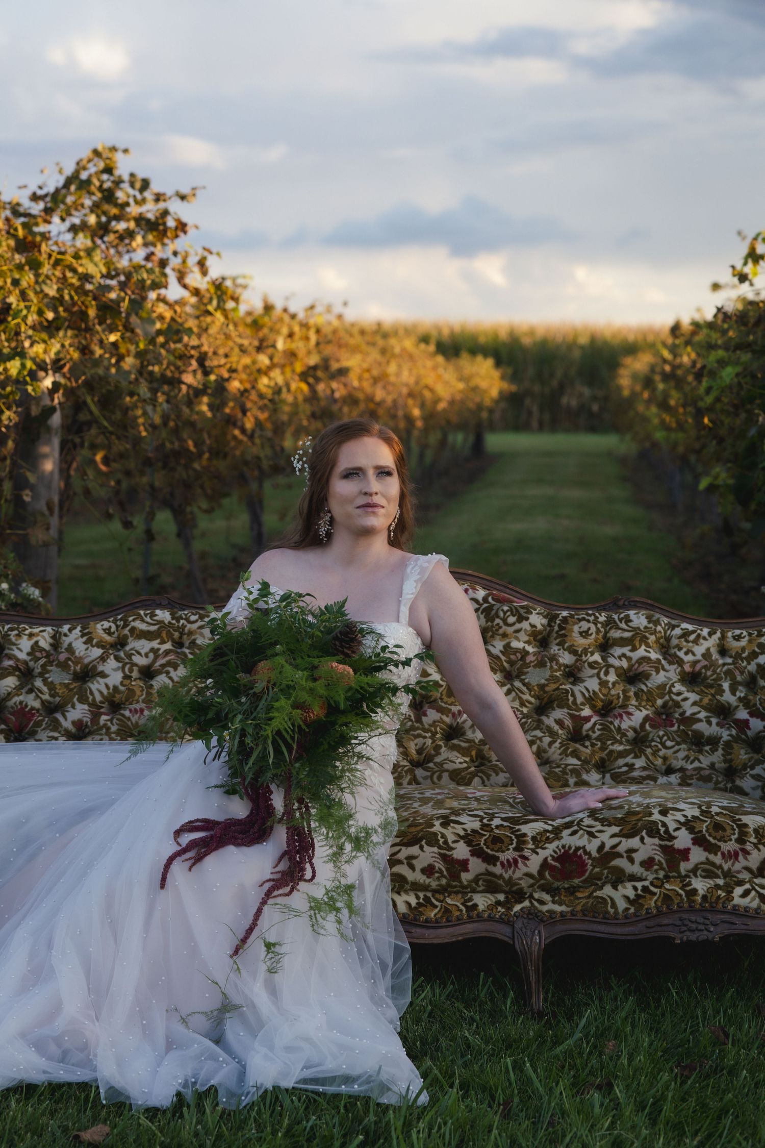 Elegant bride in white dress reclining on vintage sofa in vineyard at golden hour.