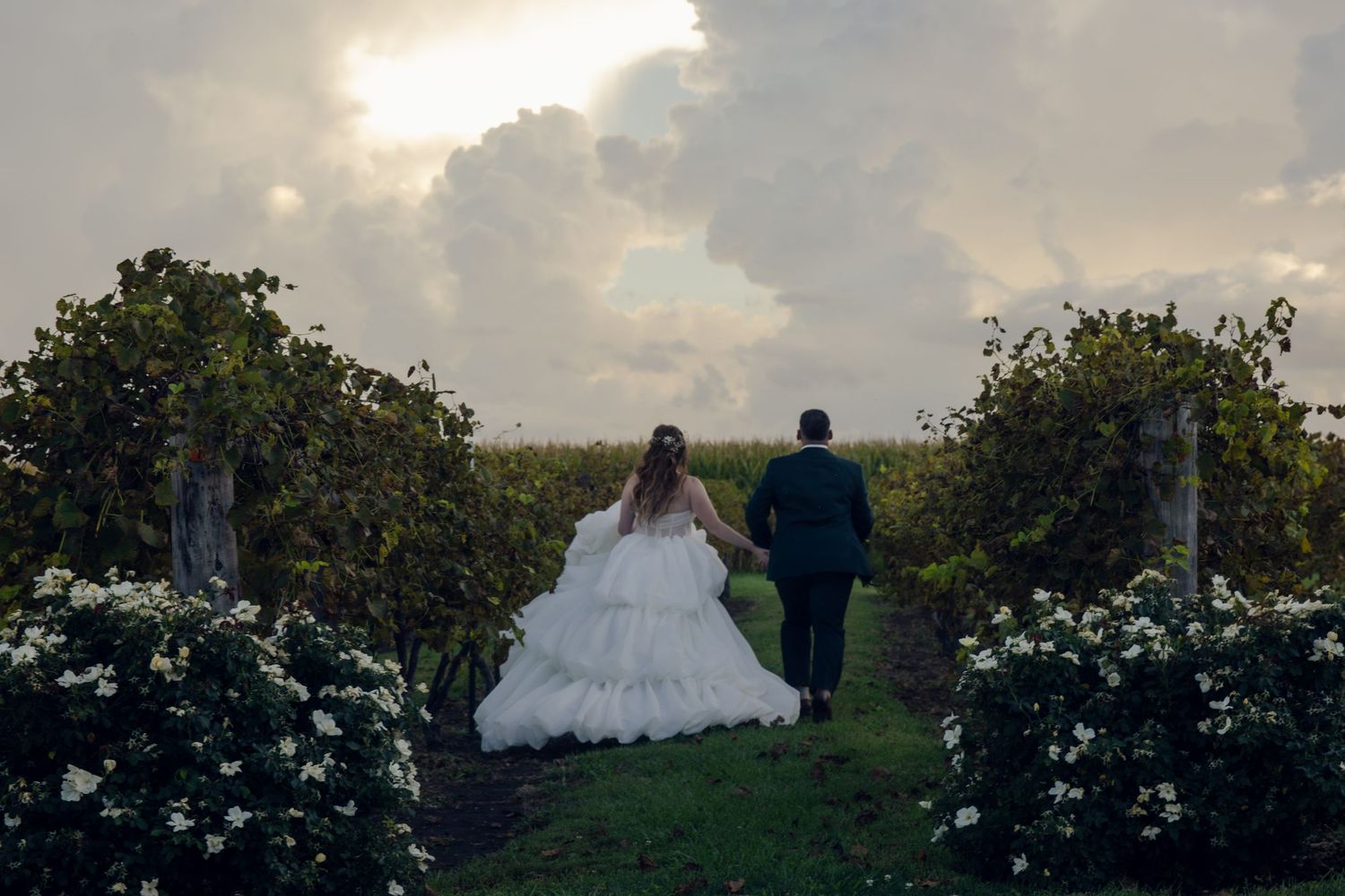Two people in wedding attire walk down a garden path lined with white flowers at sunset.