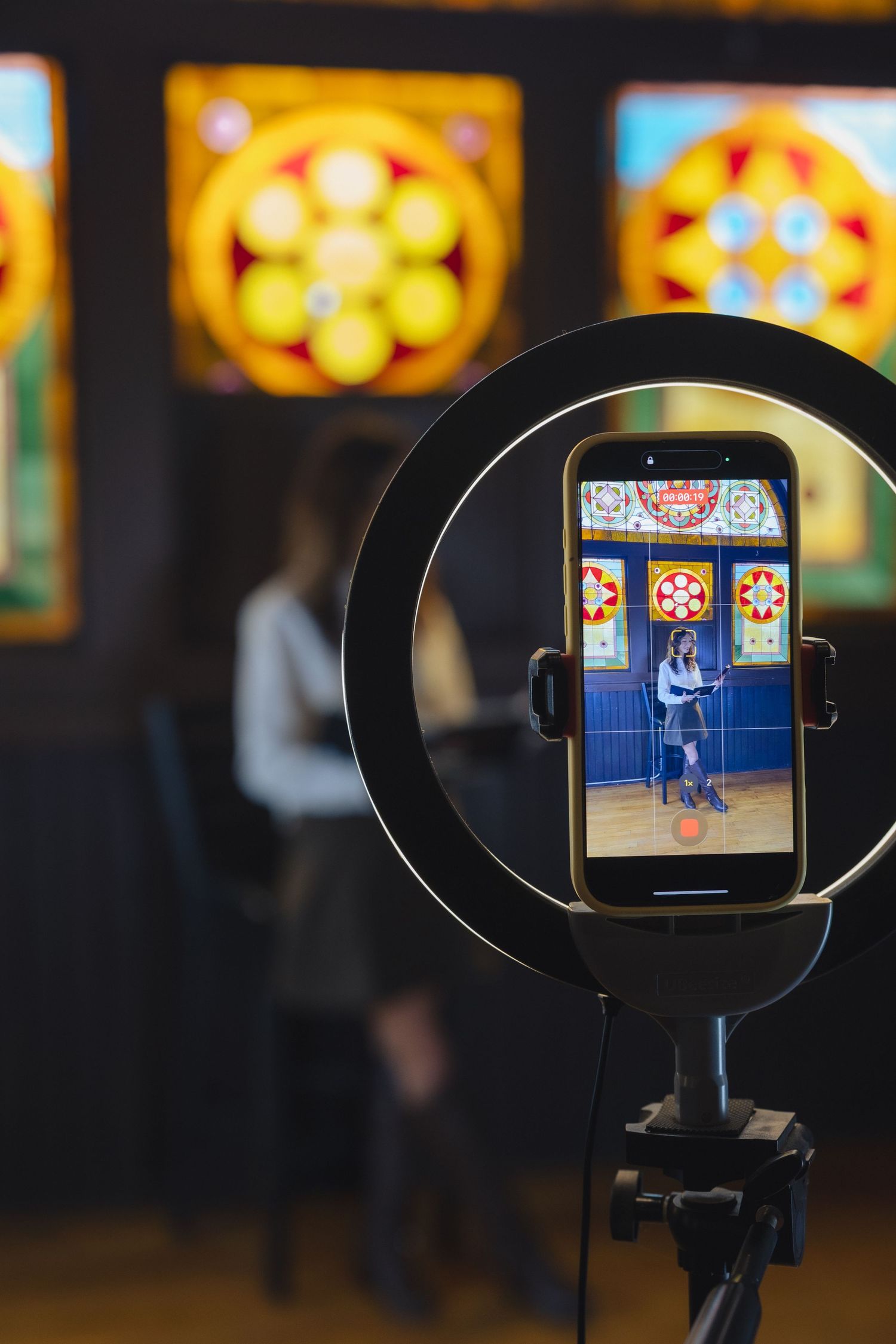 Smartphone on ring light tripod capturing woman posing before colorful stained glass windows in studio.