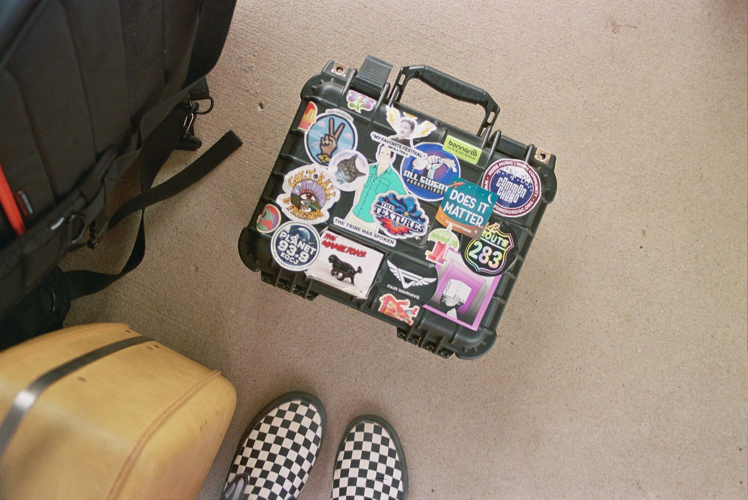 Black hard case covered in colorful travel stickers on carpet floor next to checkered Vans shoes and luggage.