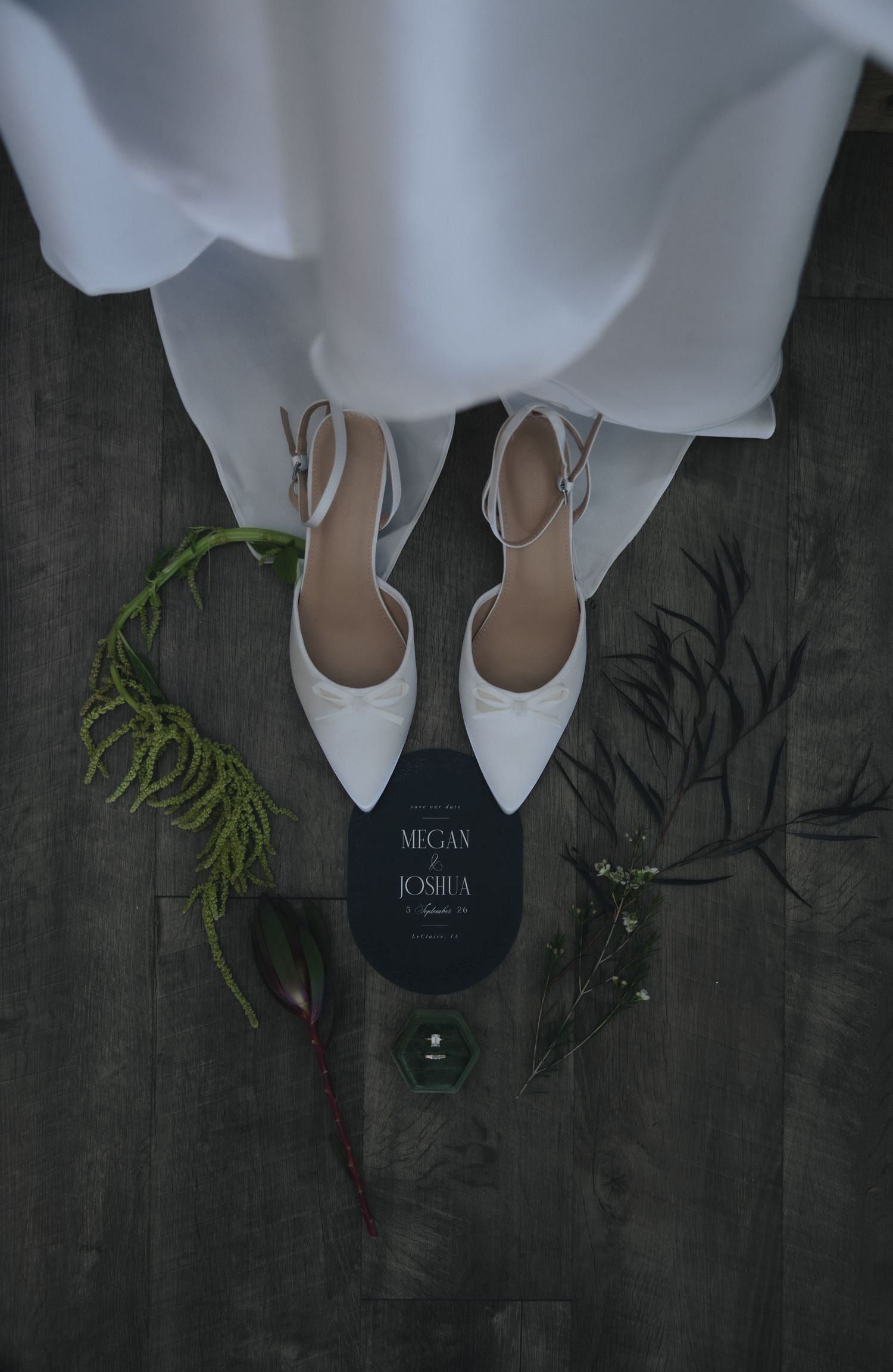 Bridal flat shoes with ankle straps on wood floor beside wedding invitation for Megan and Joshua with green botanicals.