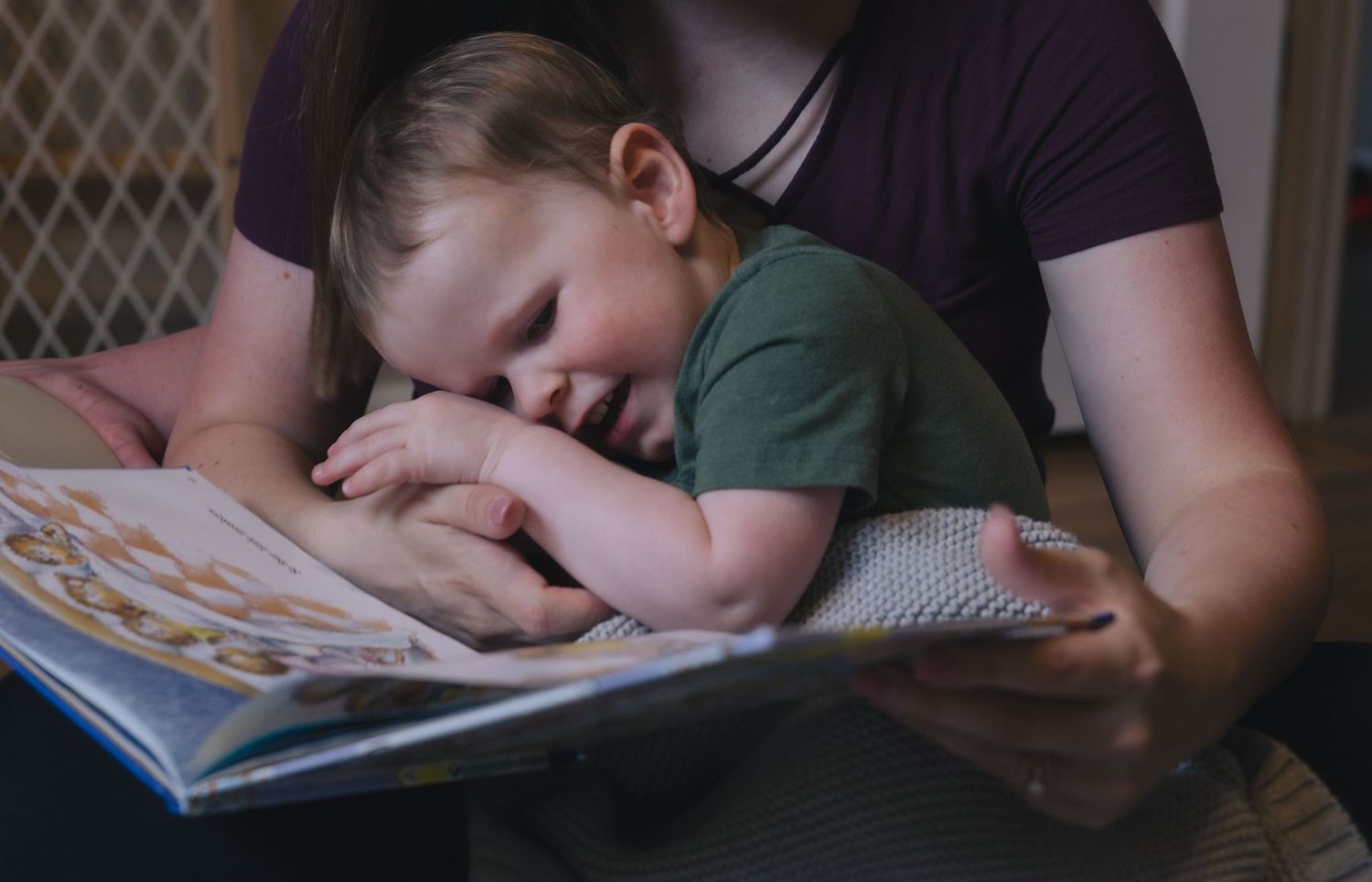 A toddler leans on an adult while reading a colorful picture book together in a cozy, warmly lit setting.