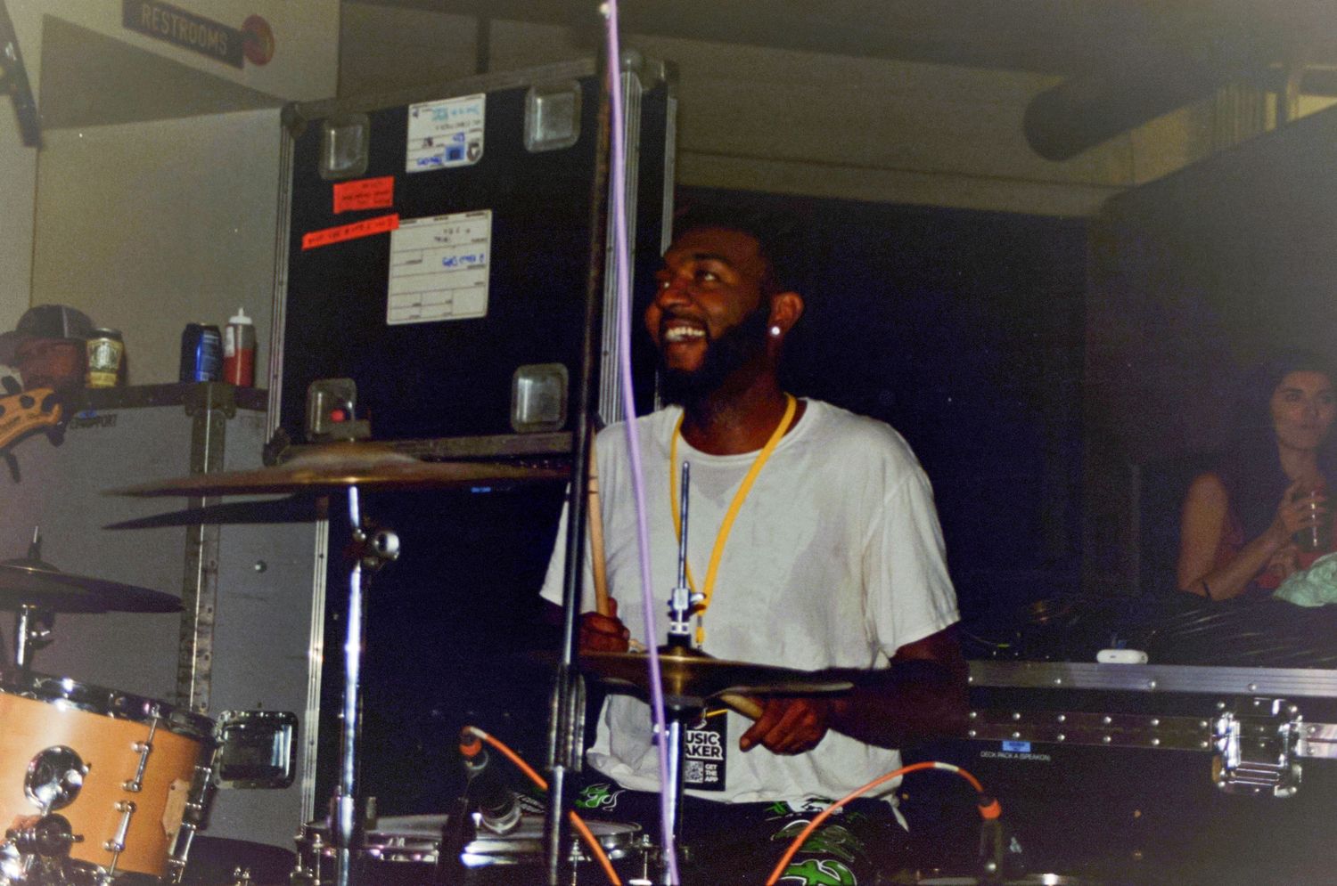 Smiling drummer in a white t-shirt playing cymbals in a dimly lit music venue.