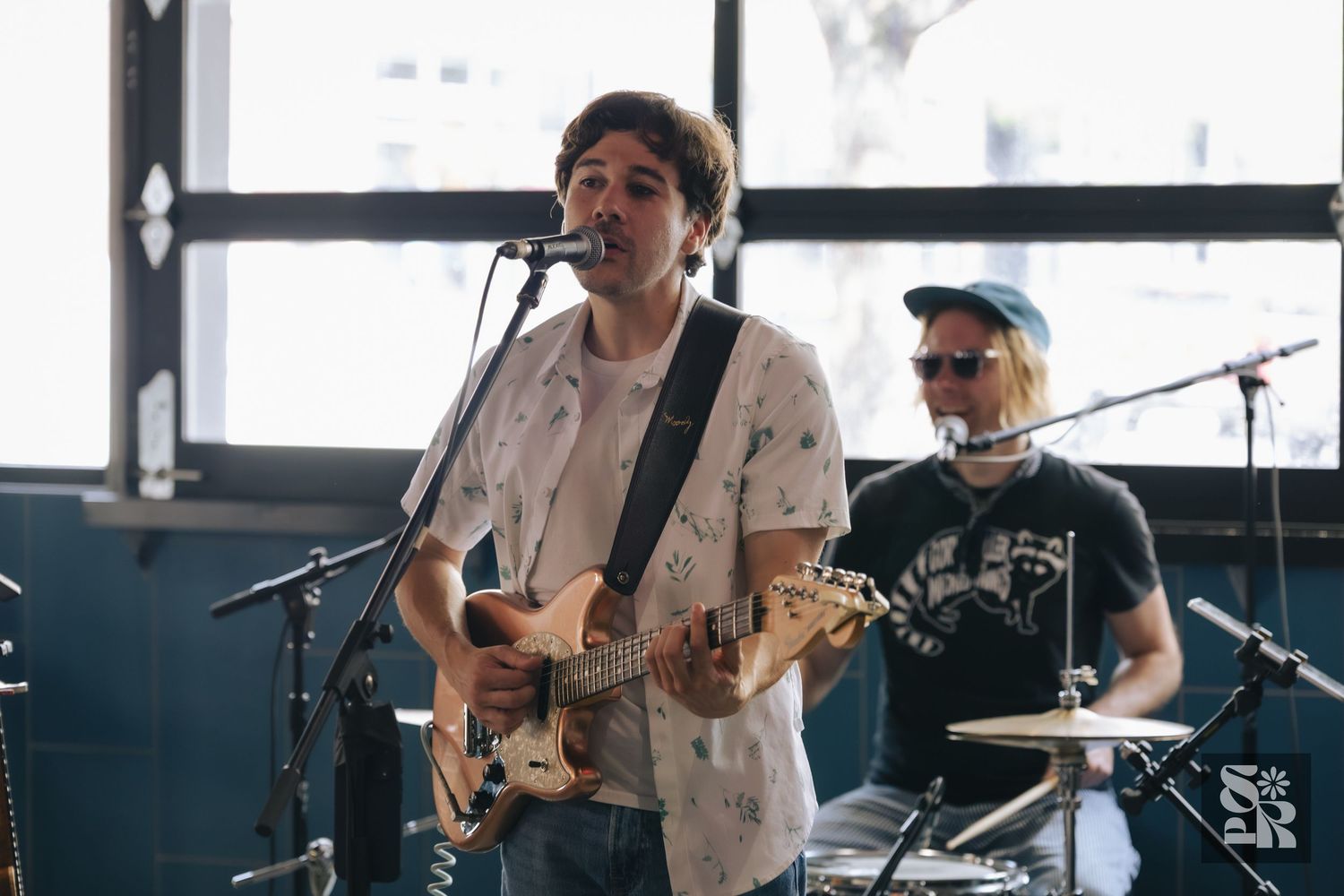 Young guitarist singing into microphone on stage with drummer performing live at an indoor music venue.
