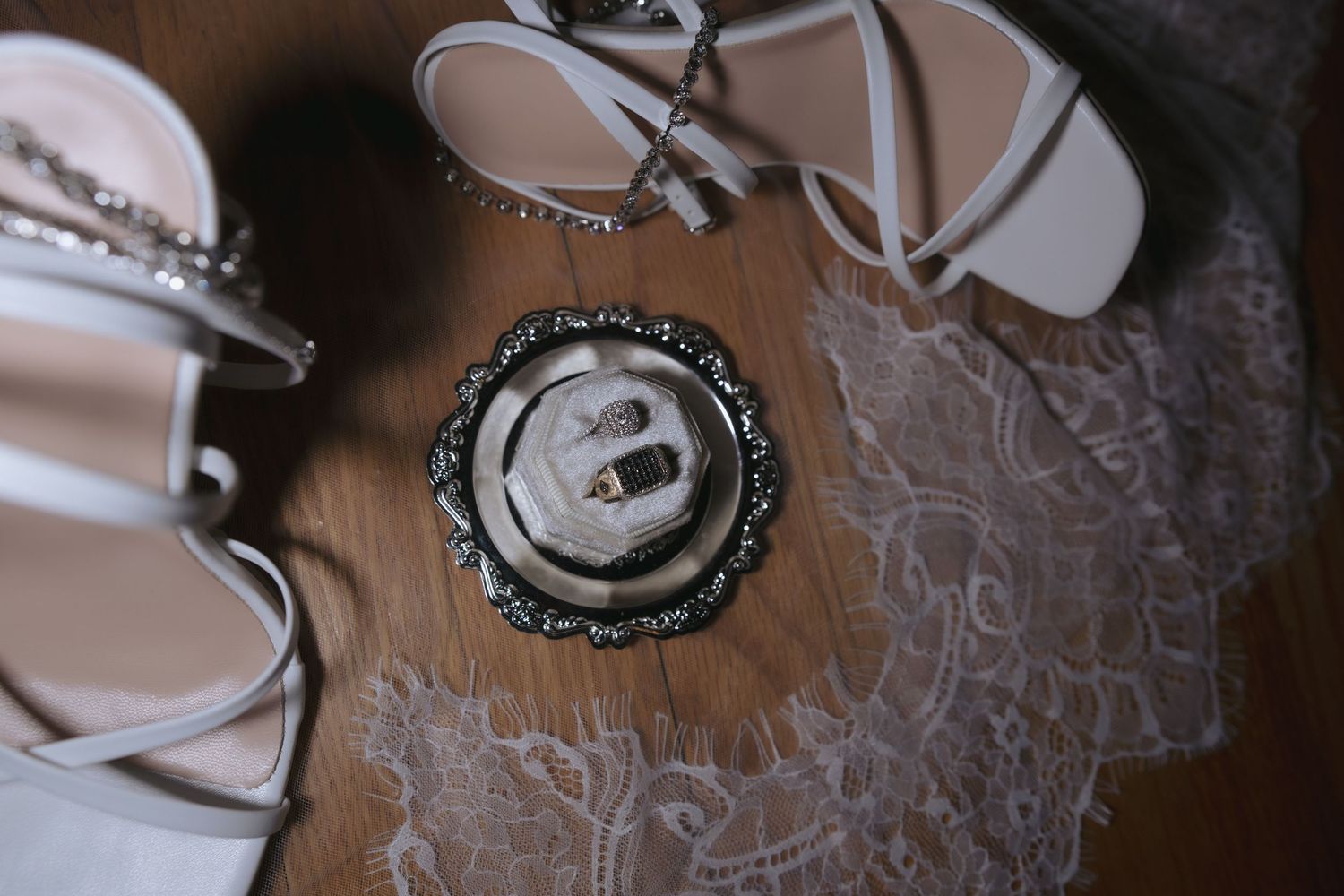 Wedding rings in ornate box beside white bridal heels and lace on wooden floor.