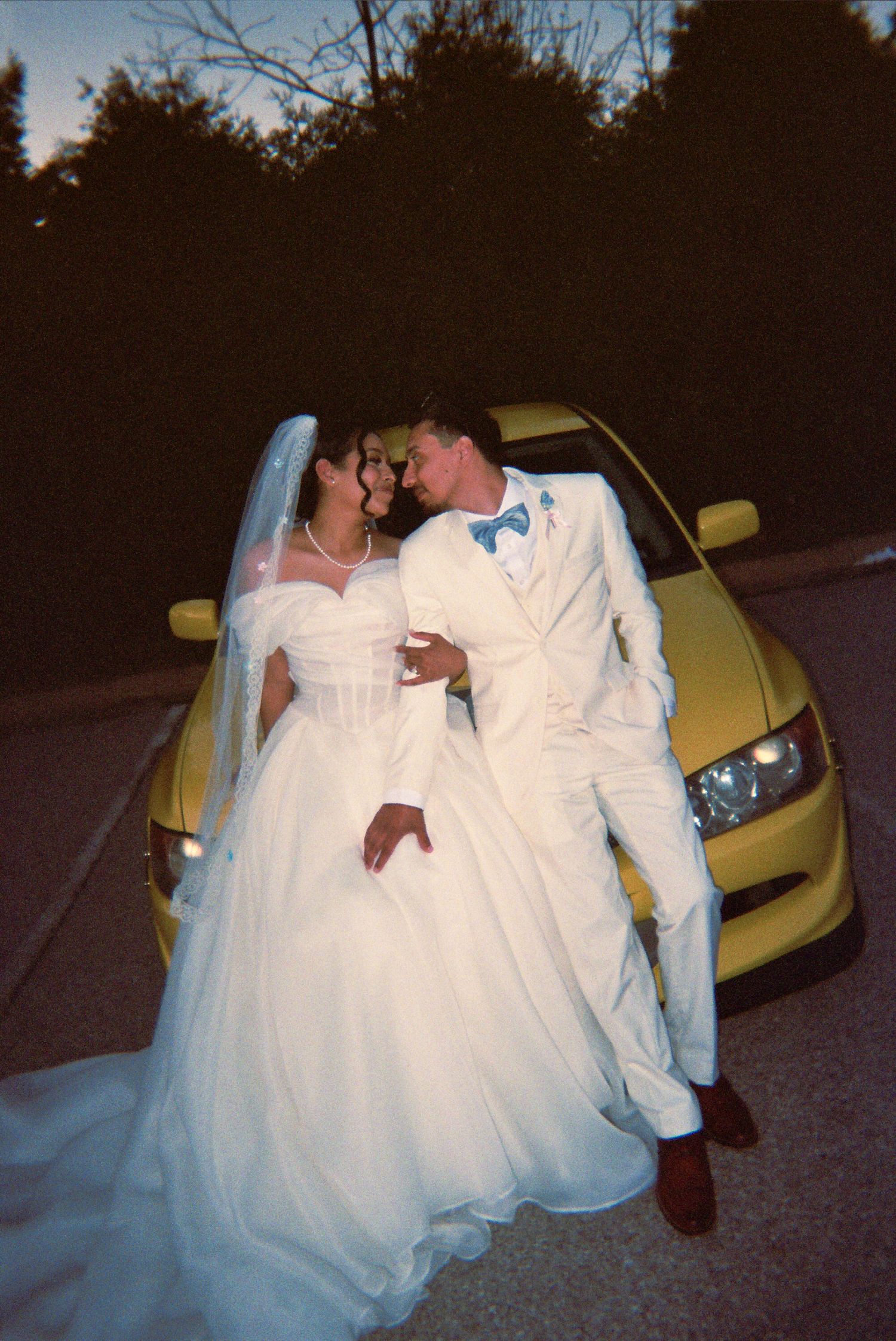 Newlyweds share a romantic moment leaning against a bright yellow car at dusk after their wedding ceremony.