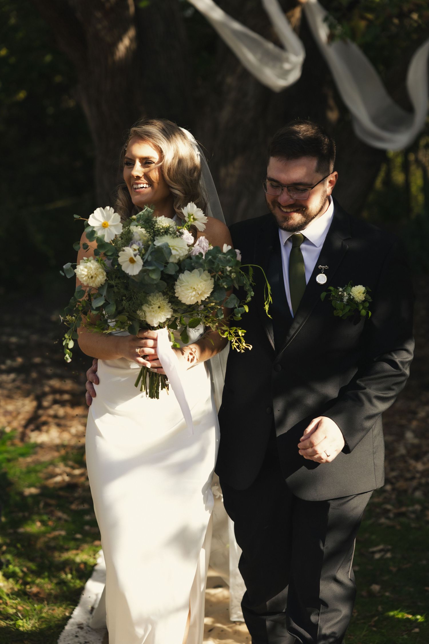 A bride in white dress holds a lush bouquet of cream and white flowers while walking with groom in black suit outdoors.