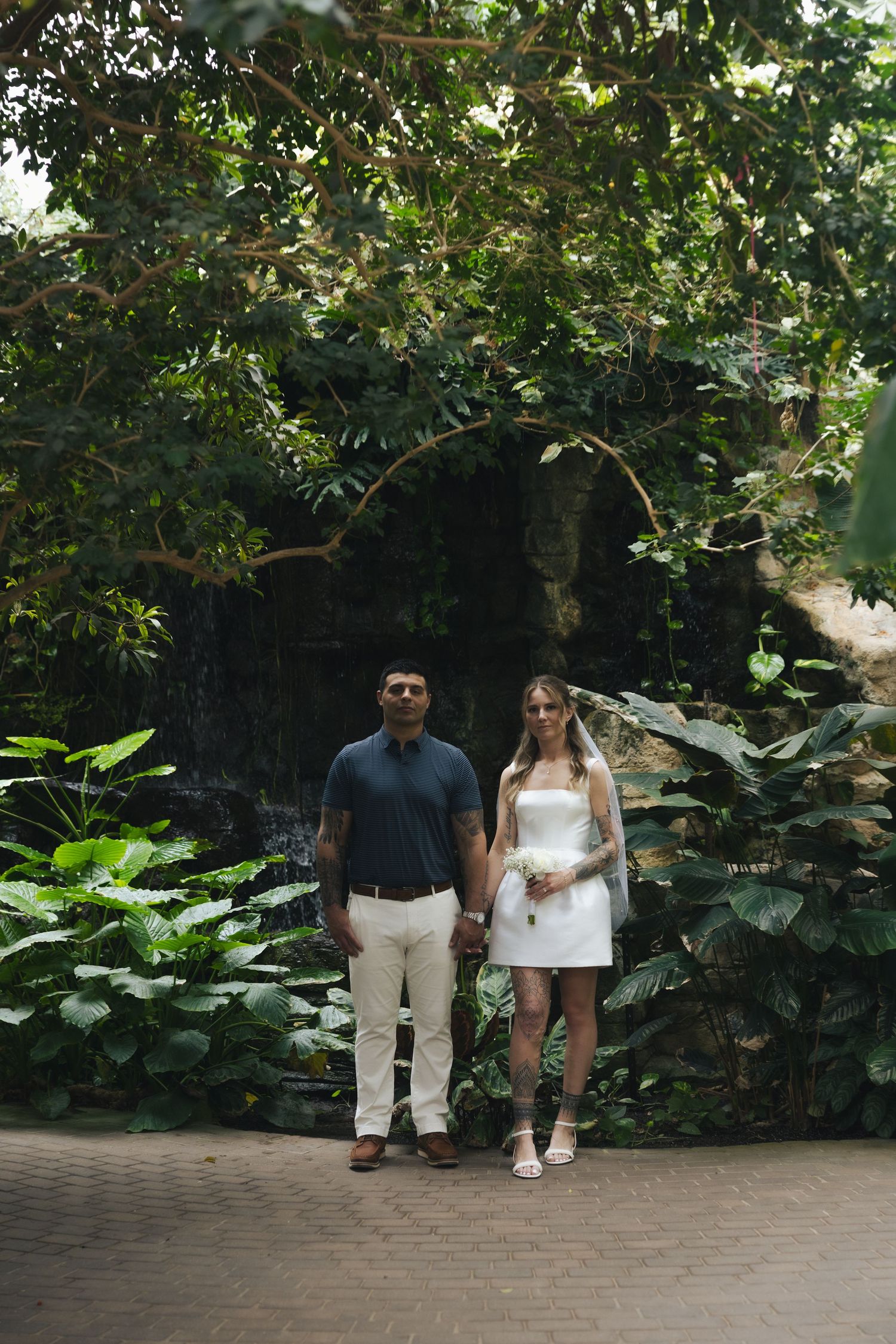 Couple in wedding attire holding hands in lush tropical greenhouse garden with waterfall backdrop.
