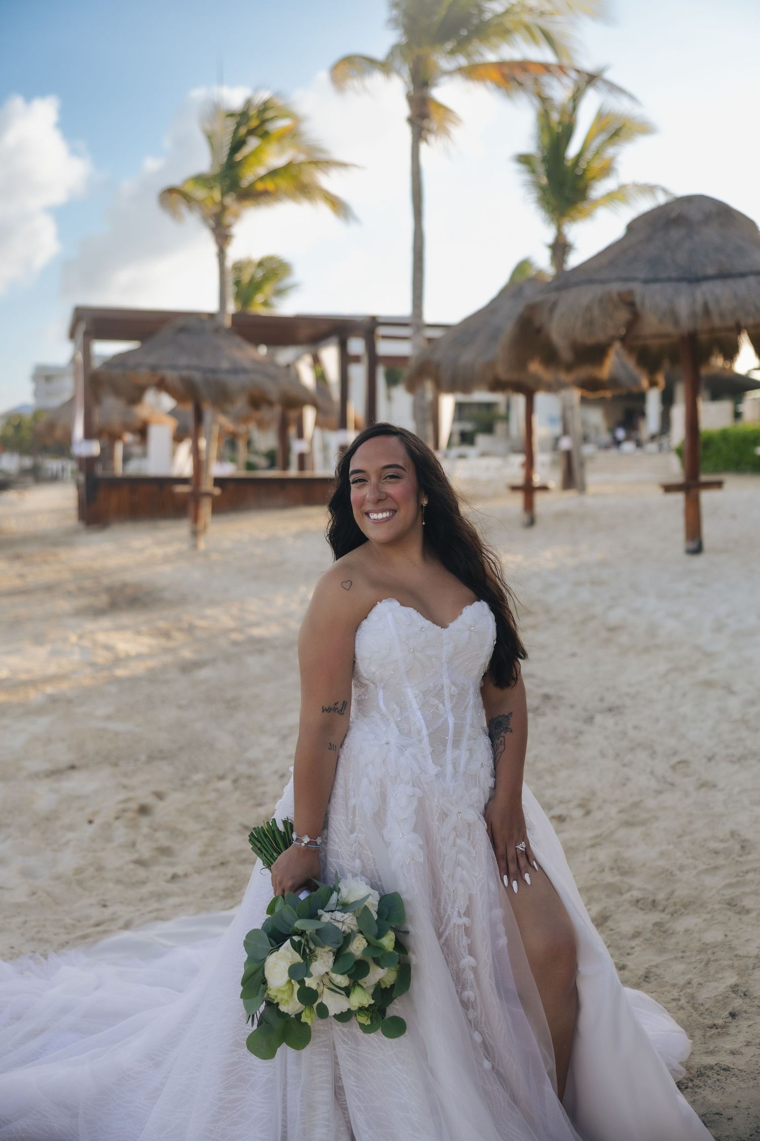 Smiling bride in strapless lace gown holding white bouquet on tropical beach with palm trees.