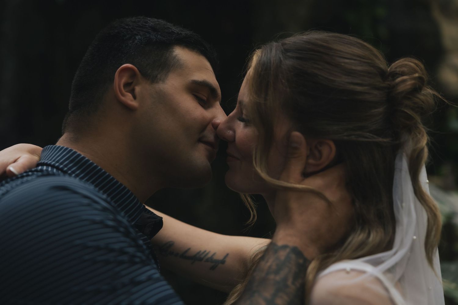 A couple with tattoos shares a romantic kiss outdoors, the woman in a white dress and man in a blue shirt.