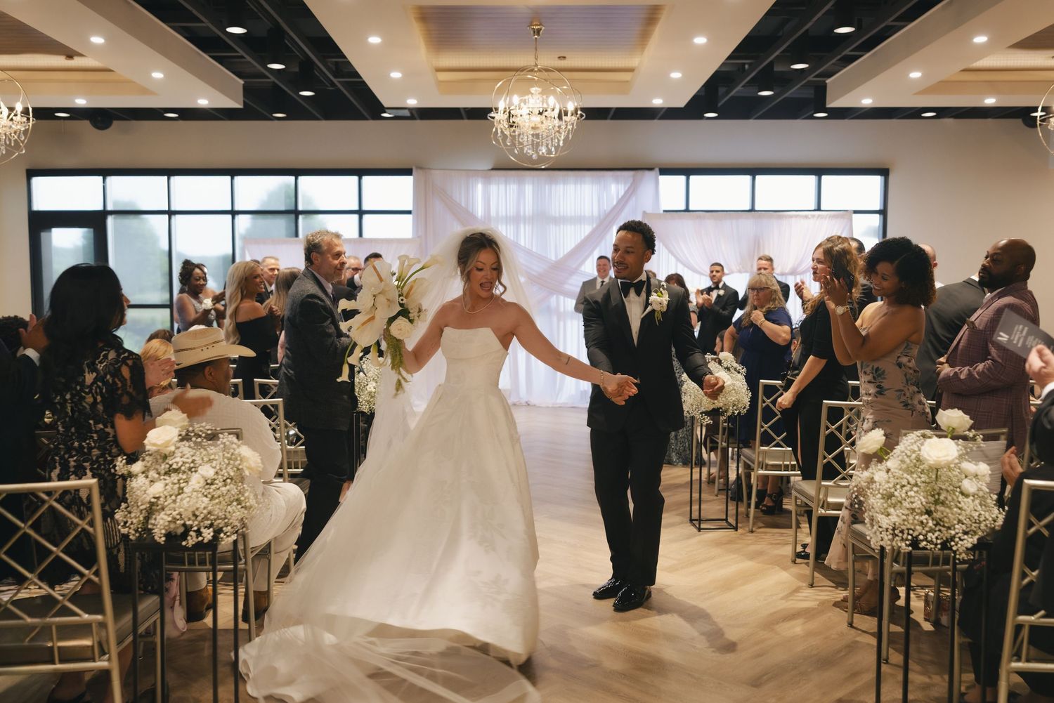 Bride and groom walk down the aisle at elegant indoor wedding ceremony with chandeliers and floral arrangements.