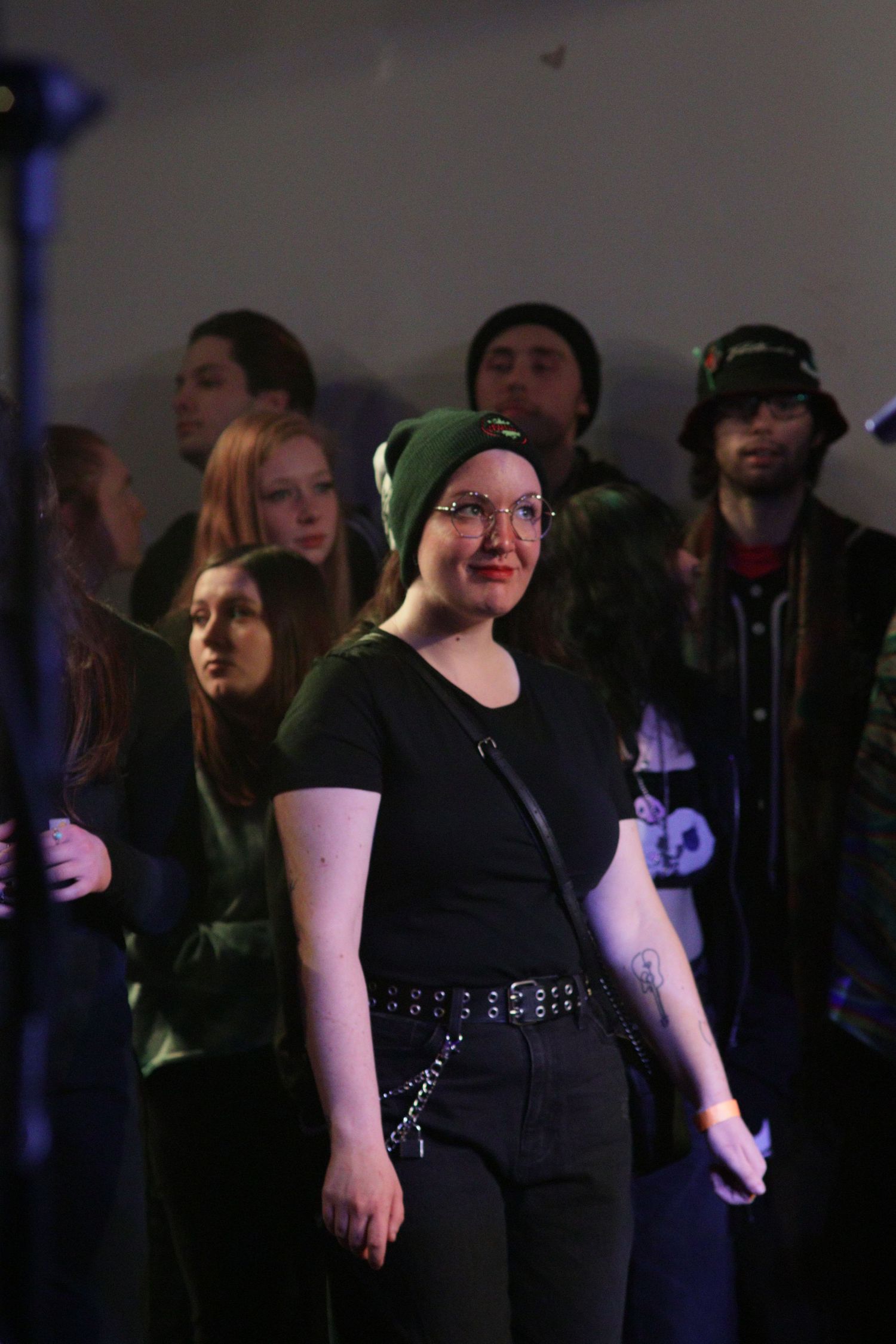 Concert audience member in black attire and beanie stands in a dimly lit venue with a chain belt accessory.